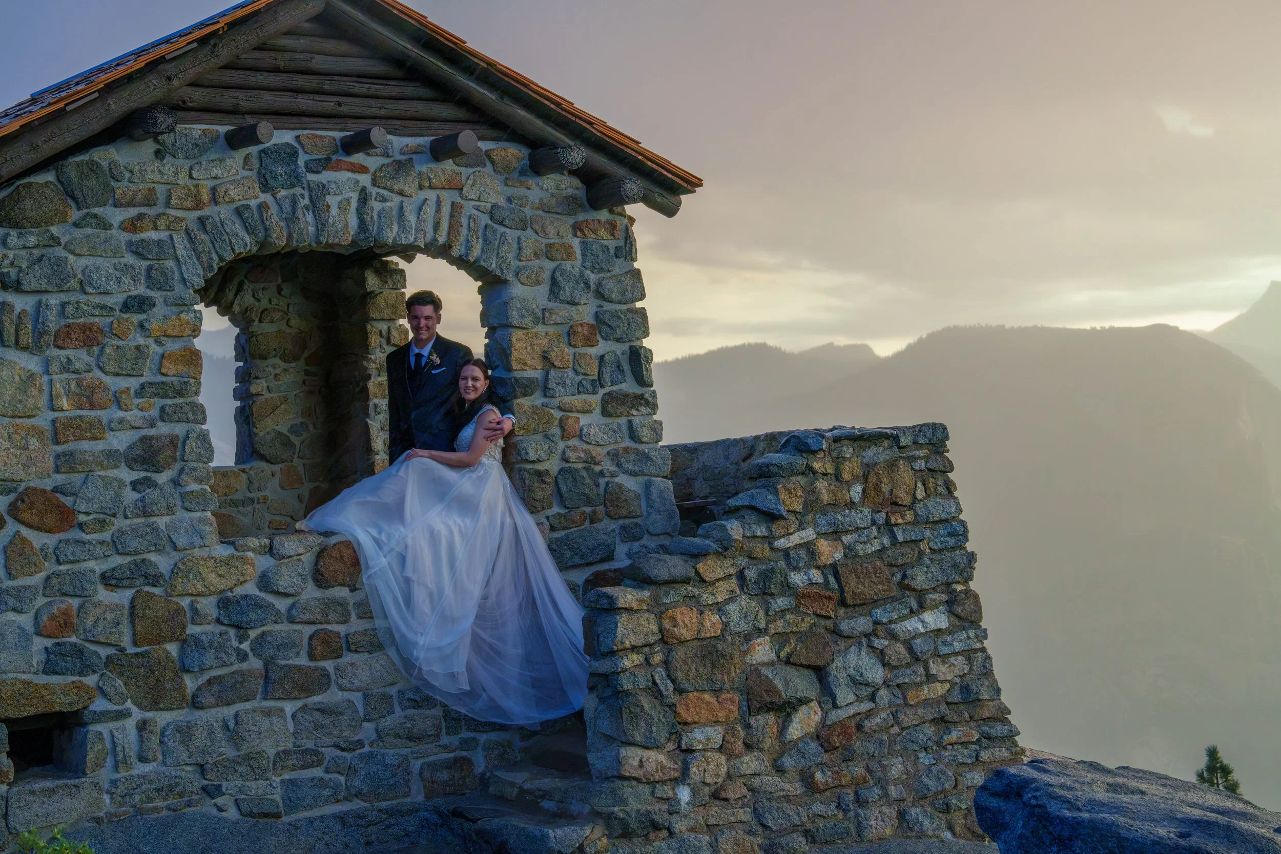 A newlywed couple, dressed in wedding attire, stands on a stone structure with a mountain and cloudy sky in the background at sunset. Yosemite Nation Park. Yosemite Elopement Photography. Yosemite Elopement Videography