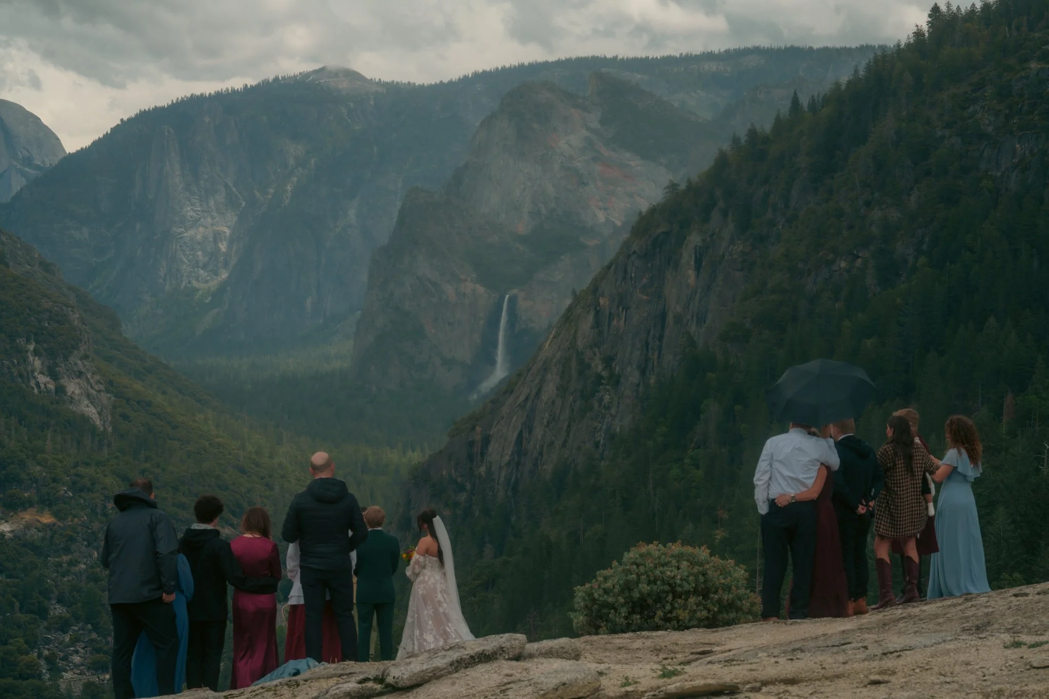People gathered outdoors on a mountain overlook with a waterfall in the background, overcast sky, during a wedding or special event.