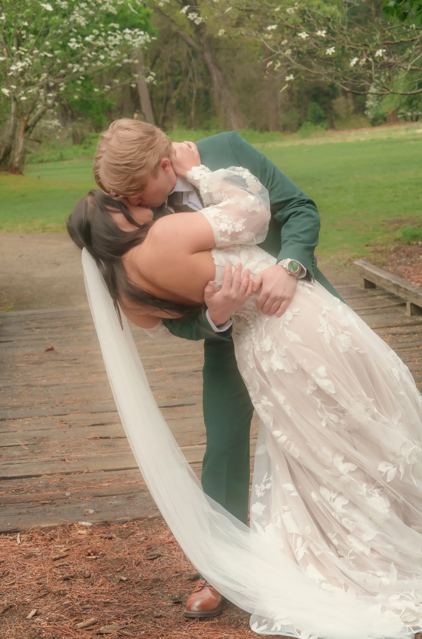 A groom in a green suit is dipping and kissing a bride in a white wedding dress with floral lace details outdoors on a wooden pathway with greenery and flowering trees in the background.