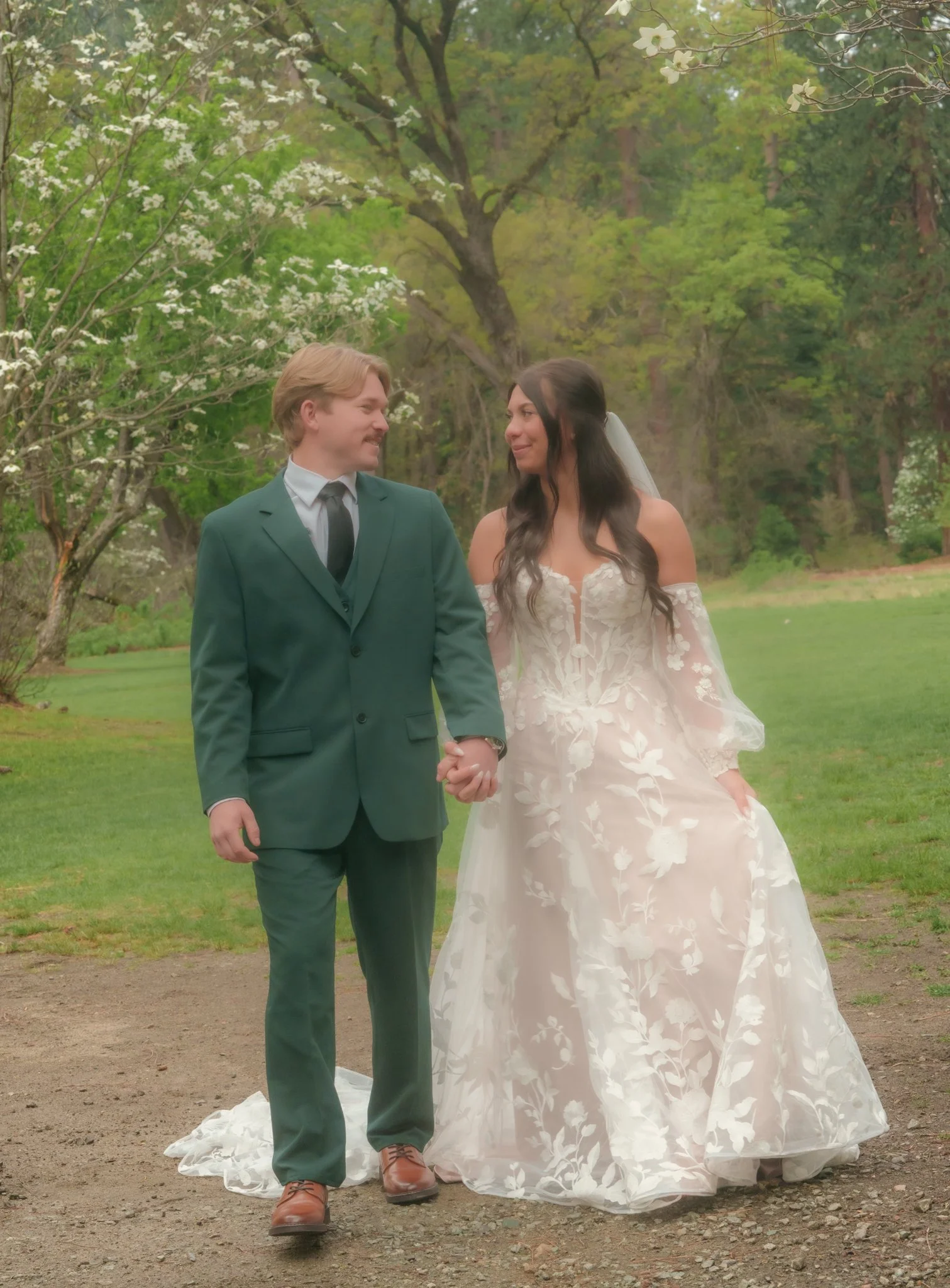 A newlywed couple holding hands and smiling at each other outdoors in a park with lush green trees and flowering bushes. The bride is wearing an off-shoulder white wedding gown with a floral pattern, and the groom is dressed in a green suit with a wh