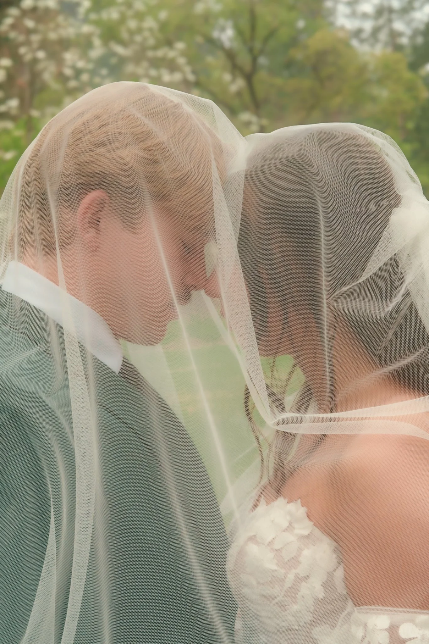 A bride and a groom wearing wedding attire under a veil, standing close together outdoors with trees in the background.