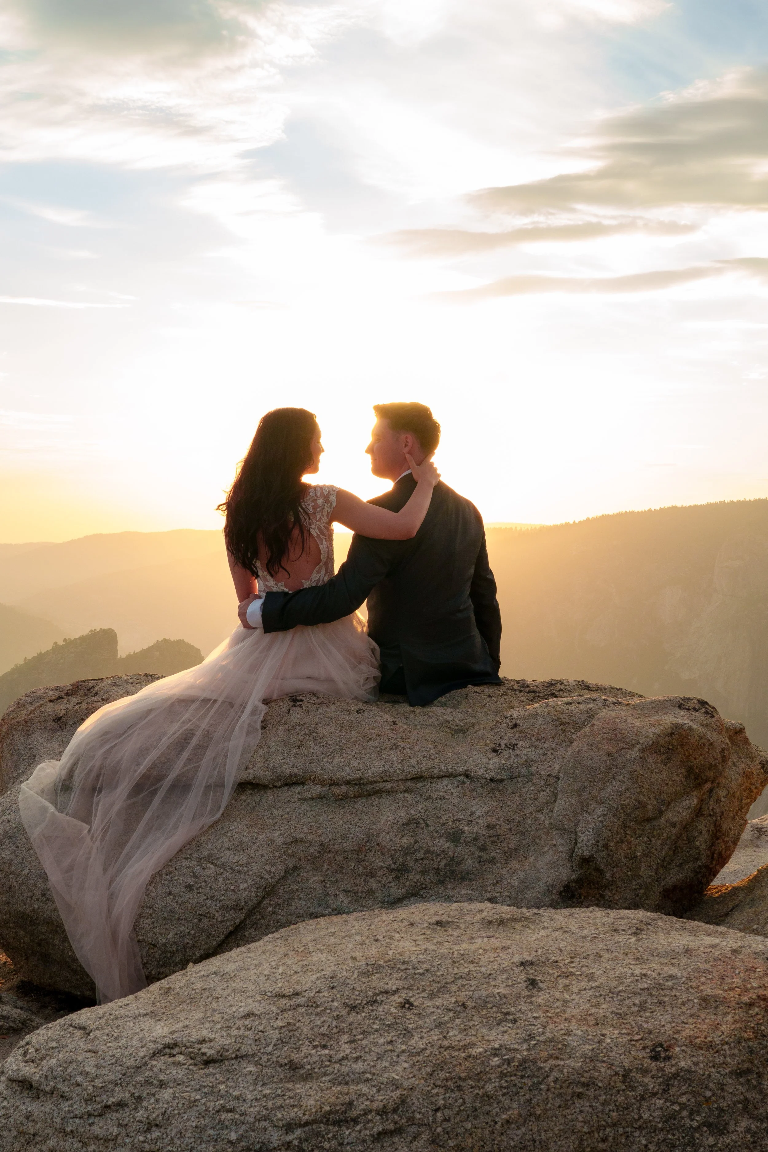 A couple dressed in wedding attire sitting on a large rock, looking at each other at sunset, with a scenic mountain landscape in the background. Yosemite Nation Park. Yosemite Elopement Photography. Yosemite Elopement Videography Taft Point