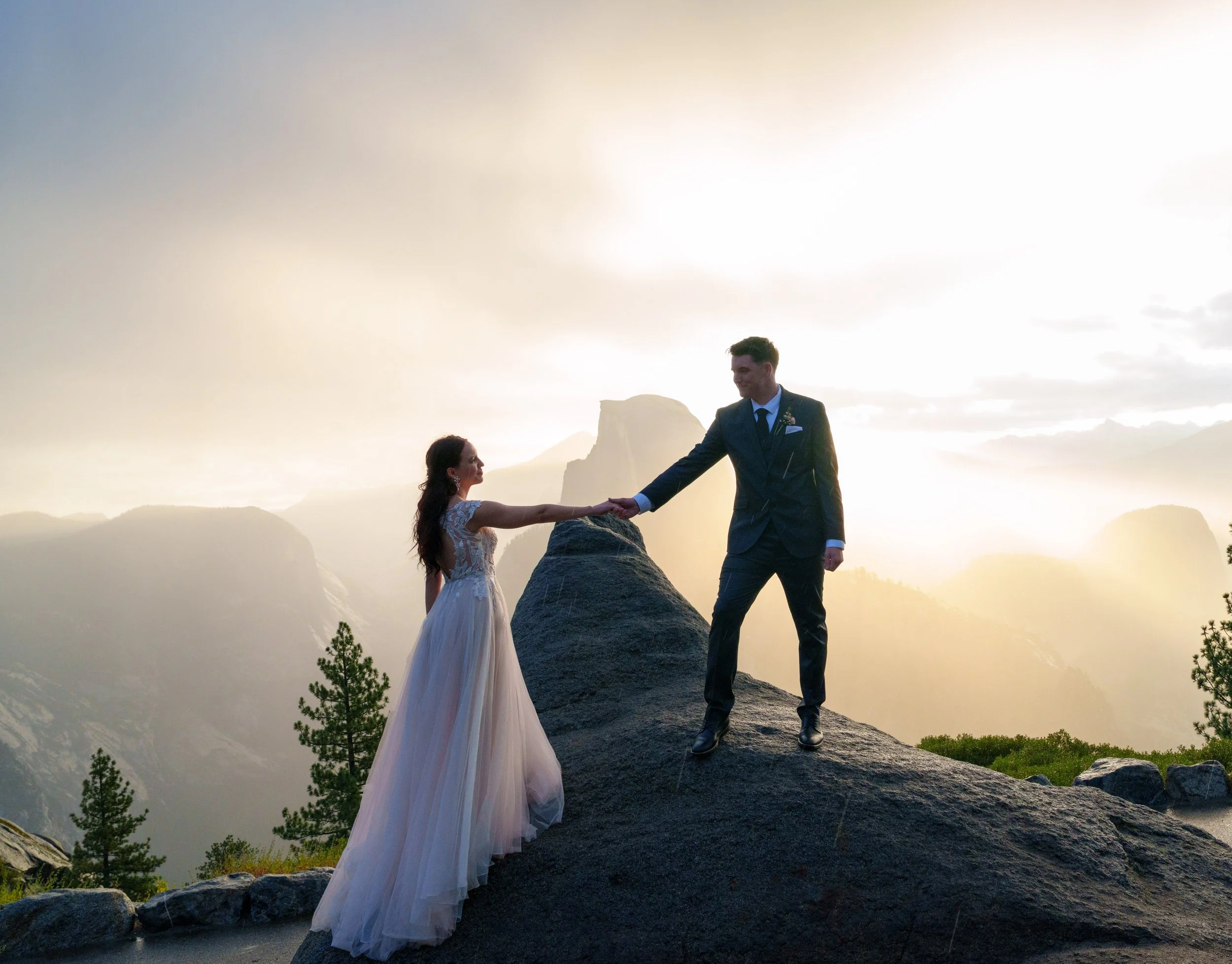 A bride and groom holding hands on a rocky ledge at sunset in a mountainous area with trees and fog. Yosemite Nation Park. Yosemite Elopement Photography. Yosemite Elopement Videography