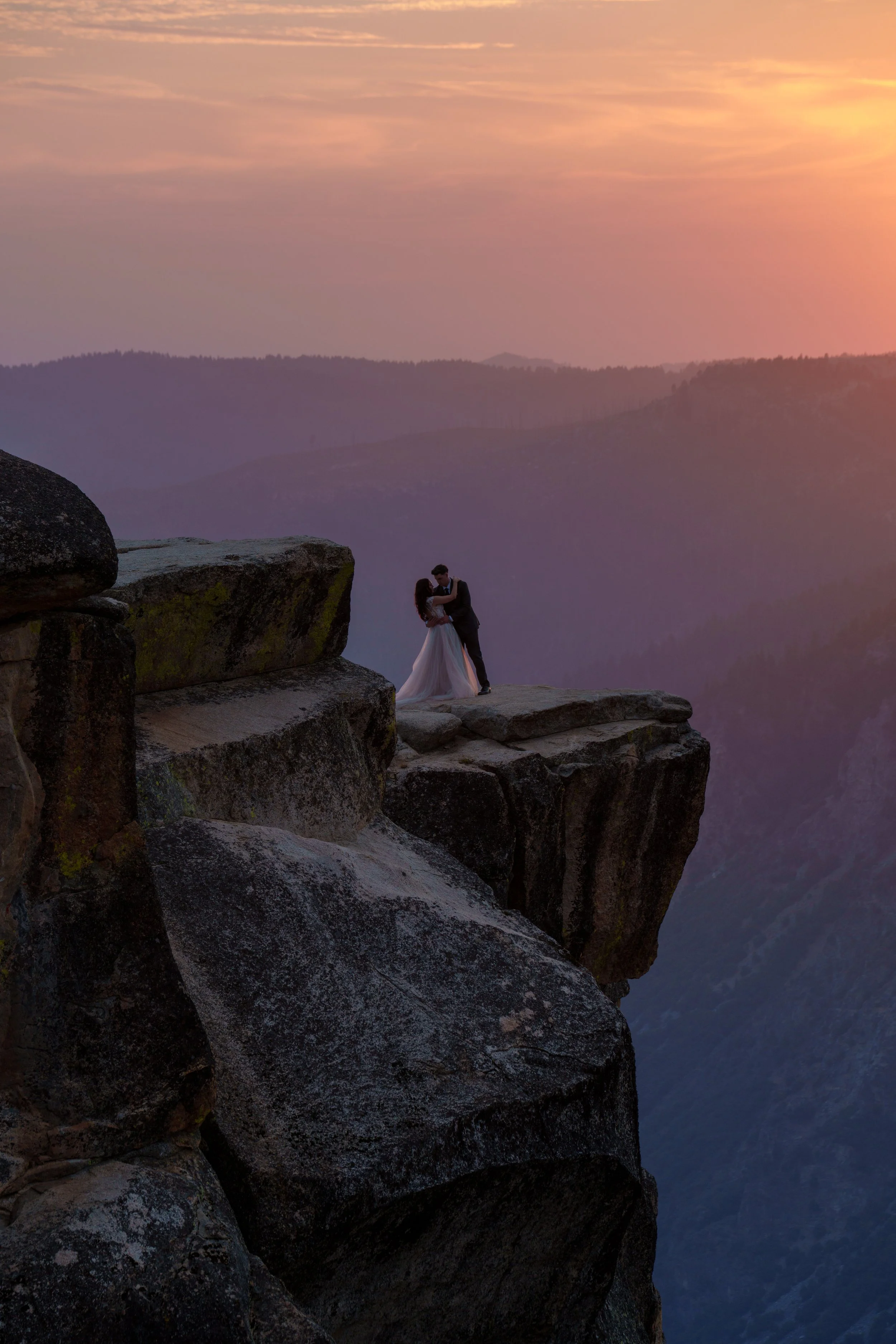 A couple in wedding attire standing on a rocky cliff at sunset, with mountains in the background and a pink, orange, and purple sky. Yosemite Nation Park. Yosemite Elopement Photography. Yosemite Elopement Videography Taft Point