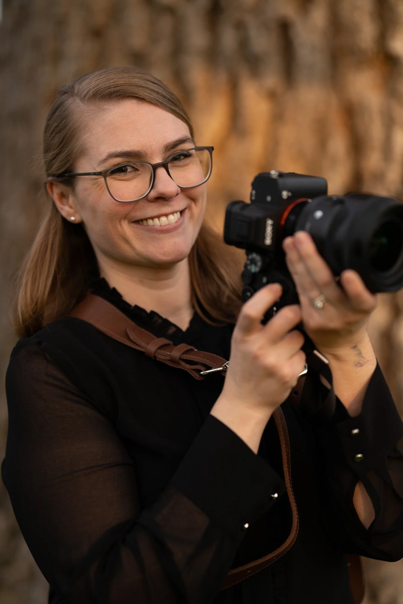 A woman with glasses smiling and holding a camera outdoors with trees in the background.