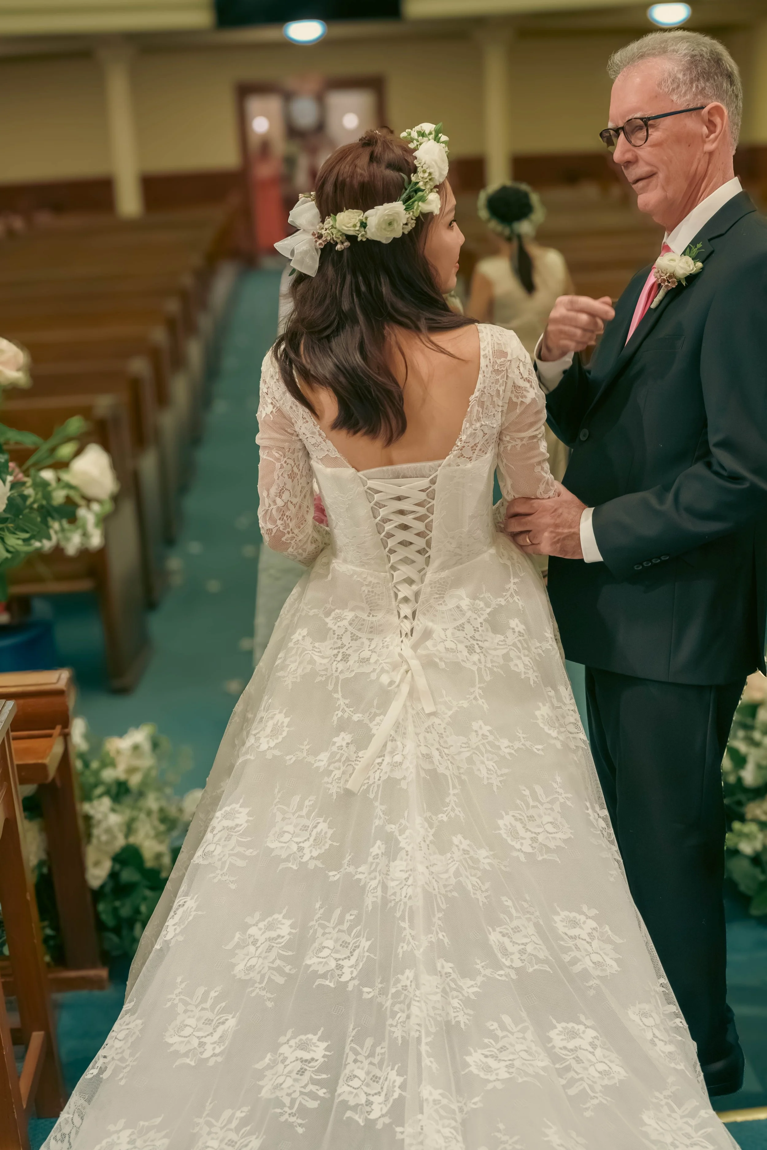 A bride in a lace wedding dress with a corset back, wearing a floral headband, standing next to an older man in a suit, in a church decorated with flowers.
