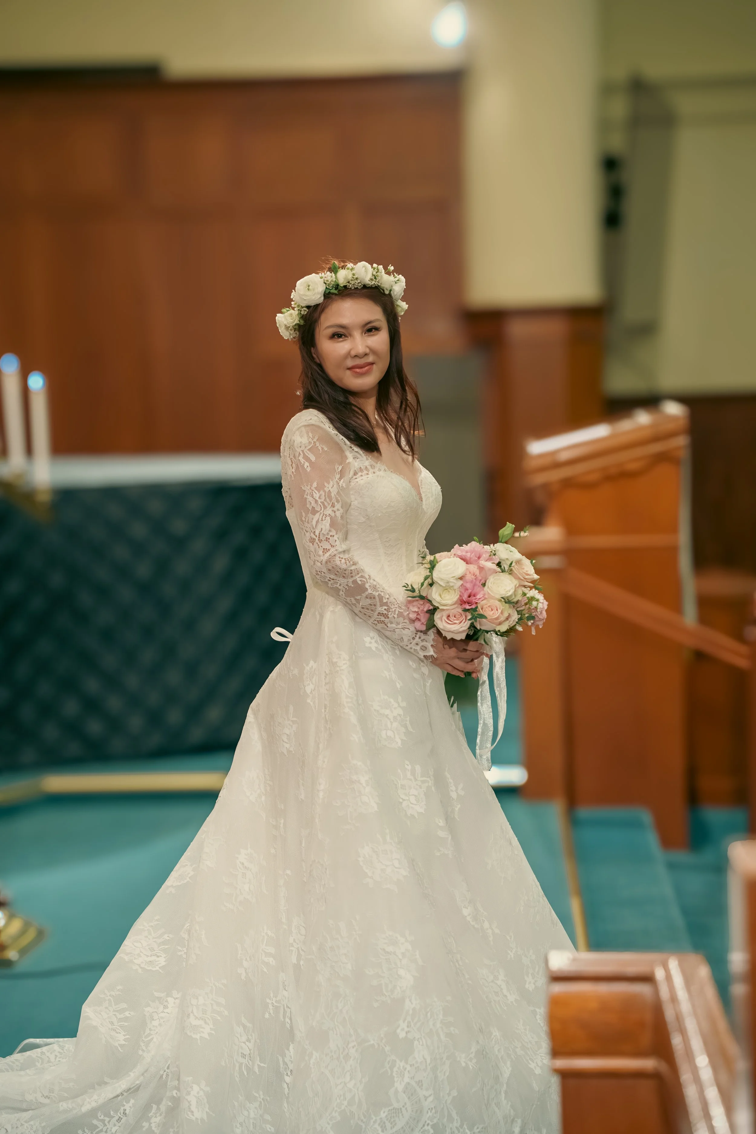A bride in a white lace wedding gown holding a bouquet of pink and white roses, wearing a floral crown, in a church or chapel setting.