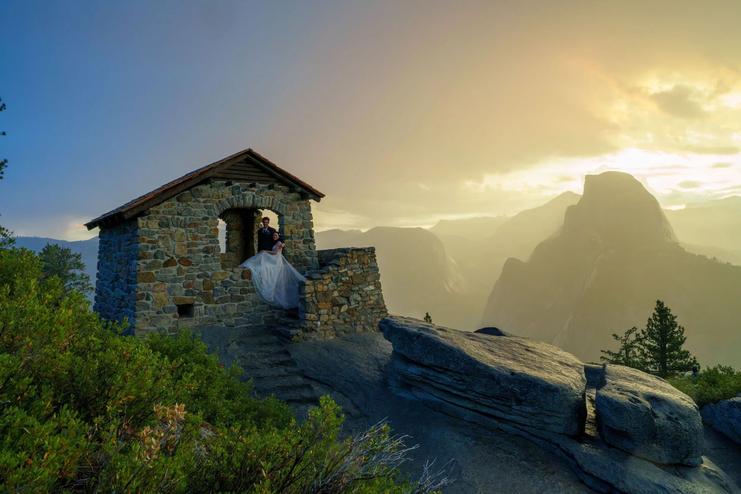 A bride and groom stand inside a small stone structure on a mountain, with misty mountain peaks in the background during sunrise or sunset. Yosemite Nation Park. Yosemite Elopement Photography. Yosemite Elopement Videography