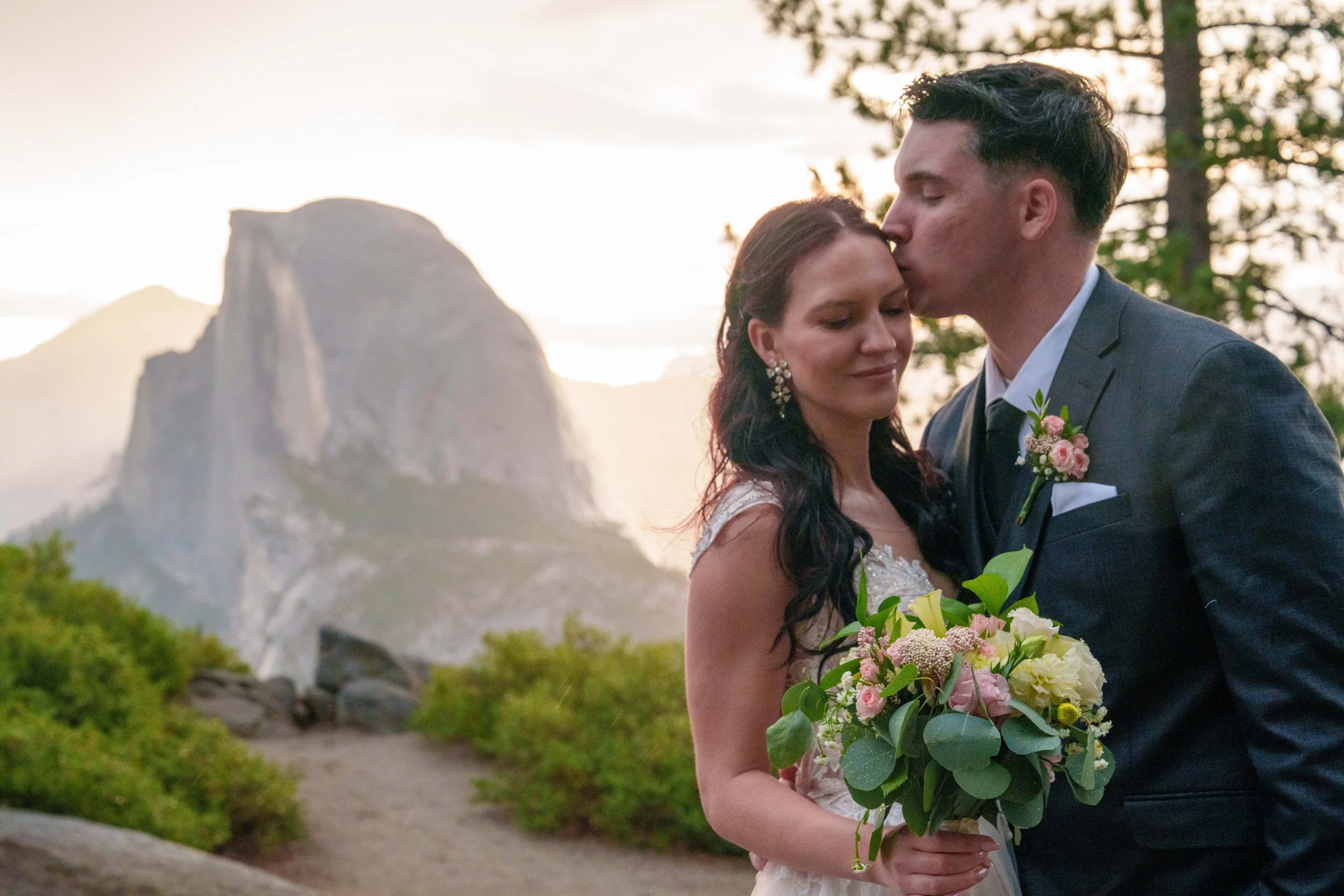 A bride and groom sharing a kiss outdoors during sunset, with a mountainous landscape and trees in the background. The bride holds a bouquet of pink, white, and yellow flowers. Yosemite Nation Park. Yosemite Elopement Photography. Yosemite Elopement 