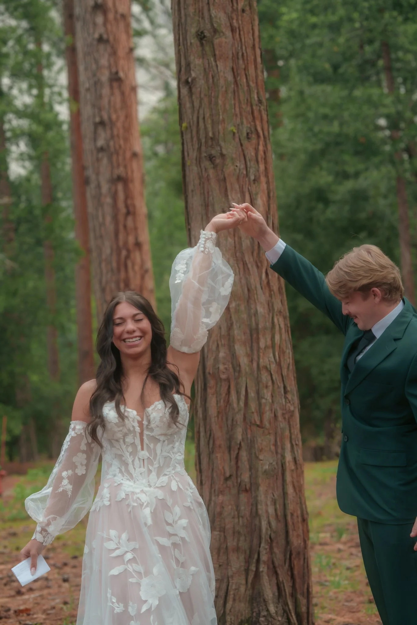 A bride in a white wedding dress, smiling, holding a piece of paper in her left hand, dancing or moving with her partner in a green suit, in a forest with tall trees.