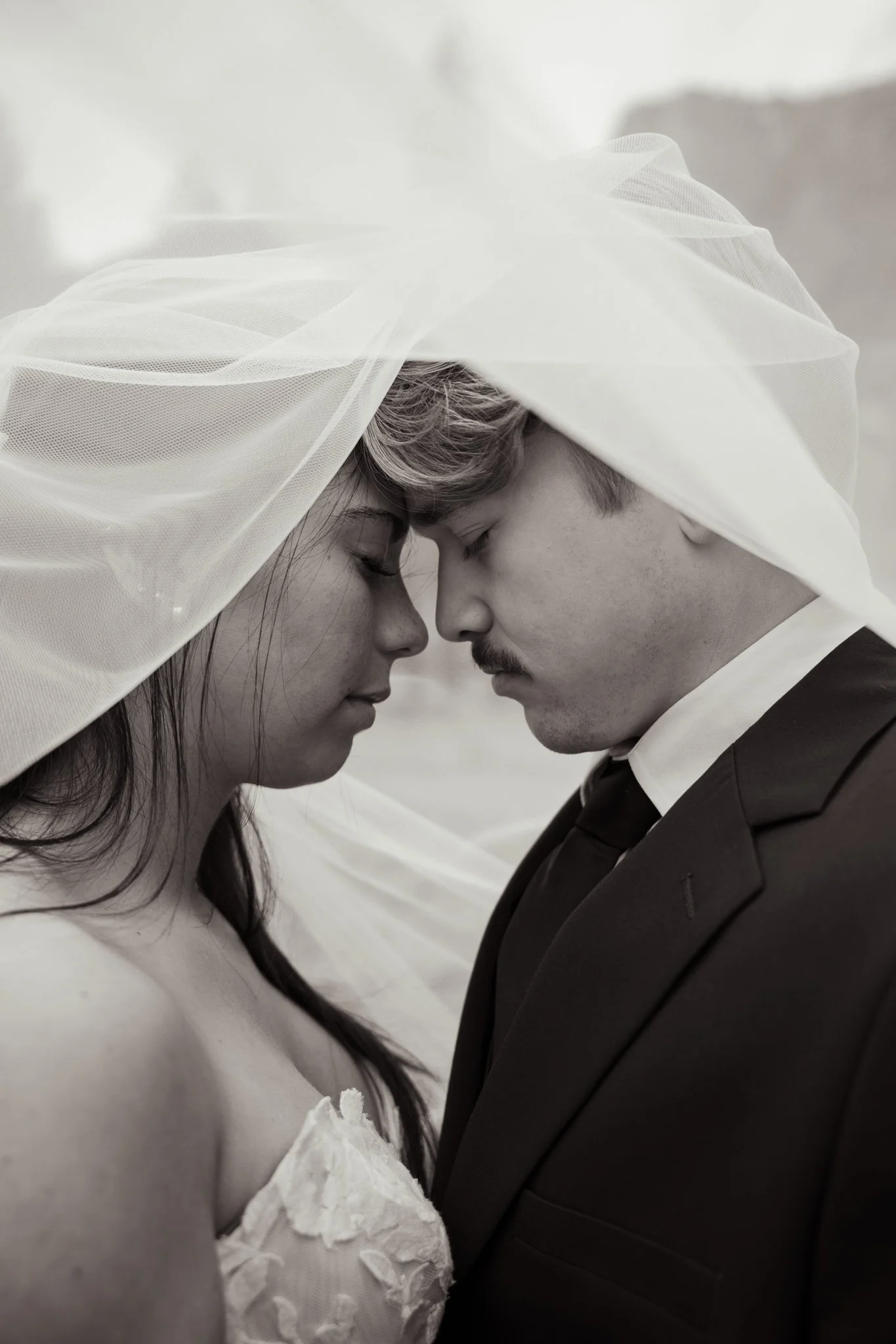 A black-and-white portrait of a couple with foreheads touching and eyes closed, wearing wedding attire with veils gently overlapping, in a romantic pose.