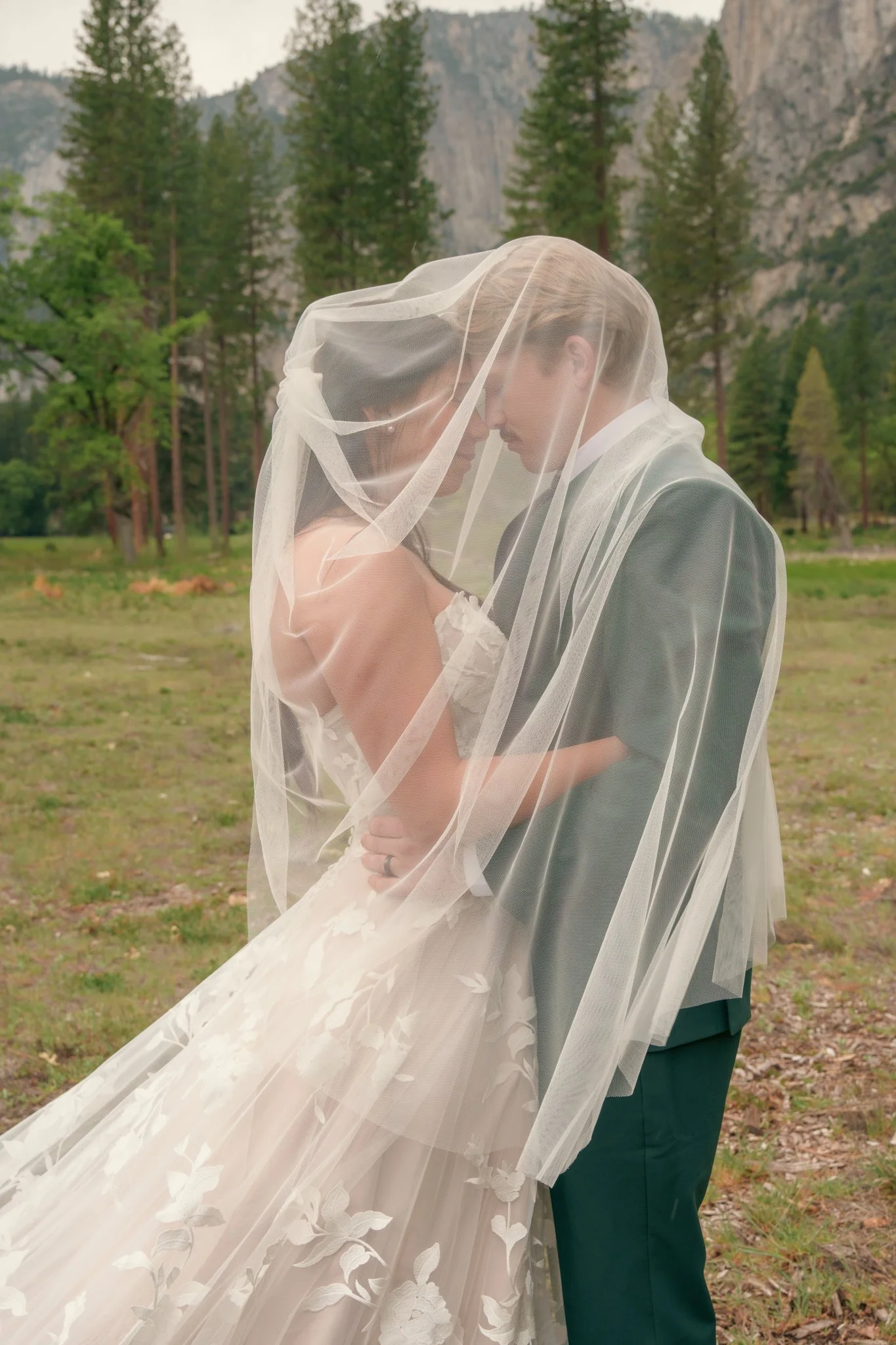 A bride and groom standing close together outdoors, wrapped in a sheer veil, with their foreheads touching and smiling. The bride is wearing a white wedding gown with floral details, and the groom is dressed in a suit. The background features a lush 