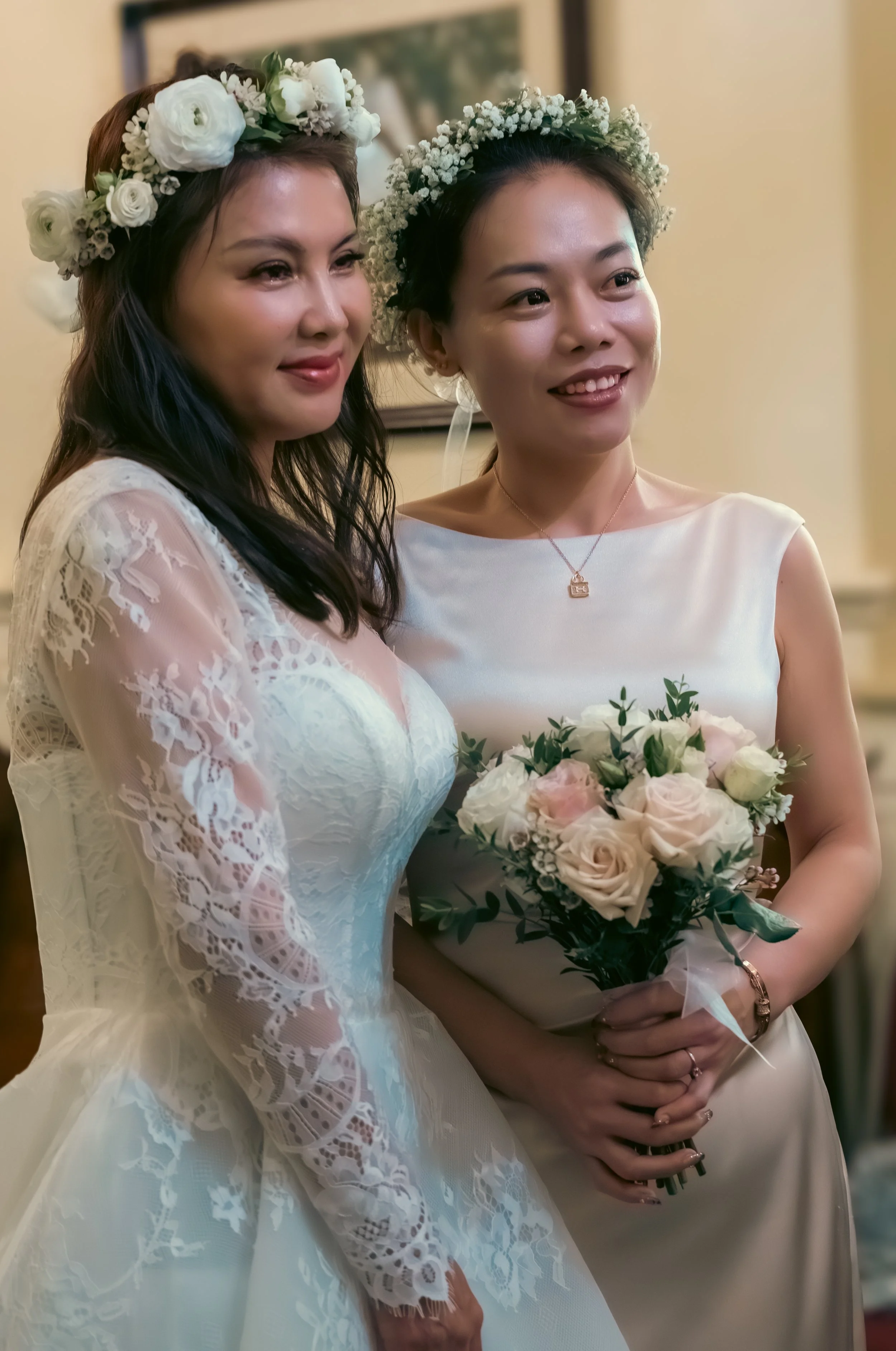 Two women dressed in wedding attire, wearing floral crowns, standing indoors. One is holding a bouquet of white and light pink roses. They are smiling and posing for a photo.