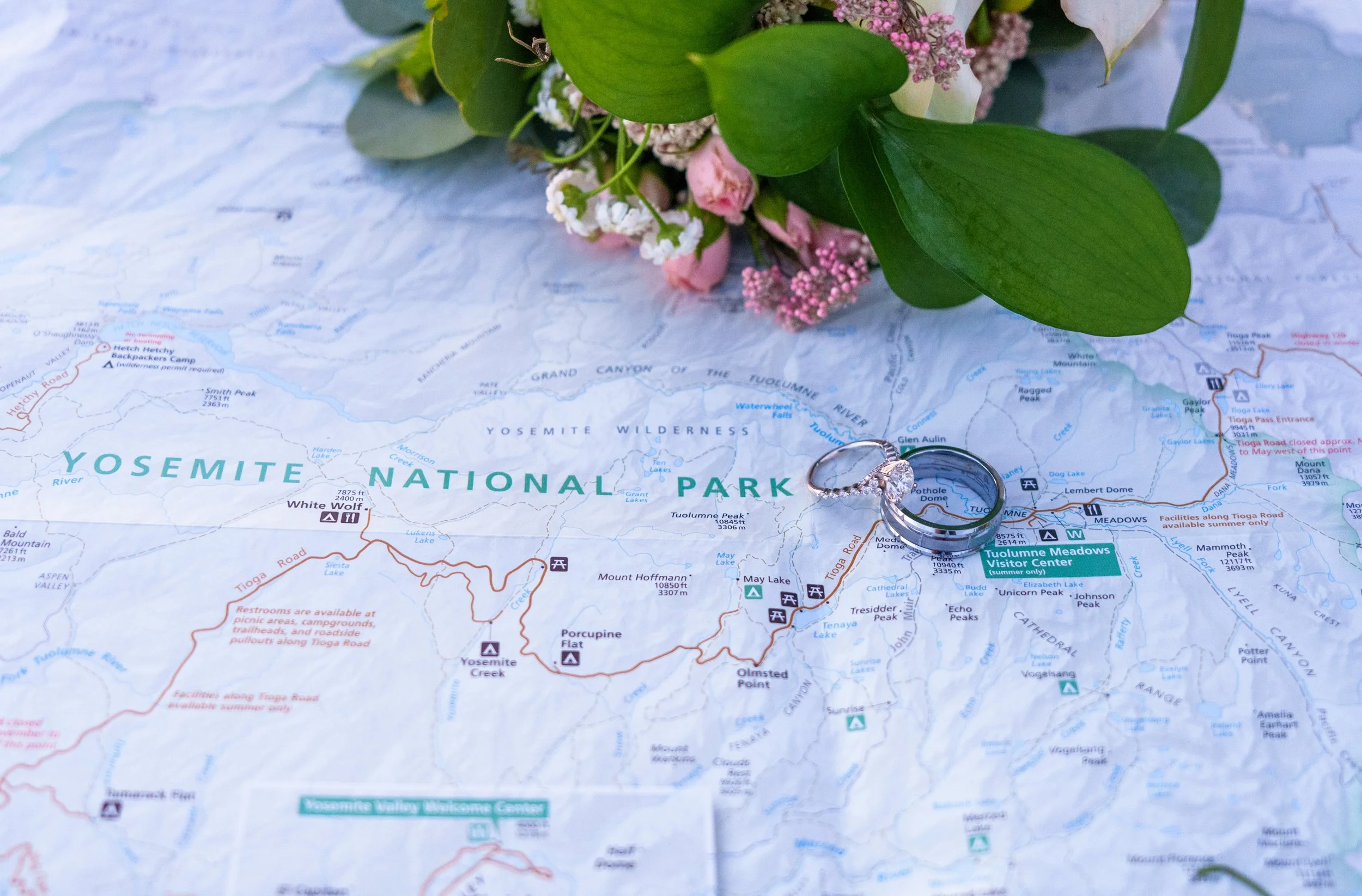 A close-up of a map of Yosemite National Park with a bouquet of pink and white flowers and green leaves resting on it, along with a silver ring. Yosemite Nation Park. Yosemite Elopement Photography. Yosemite Elopement Videography