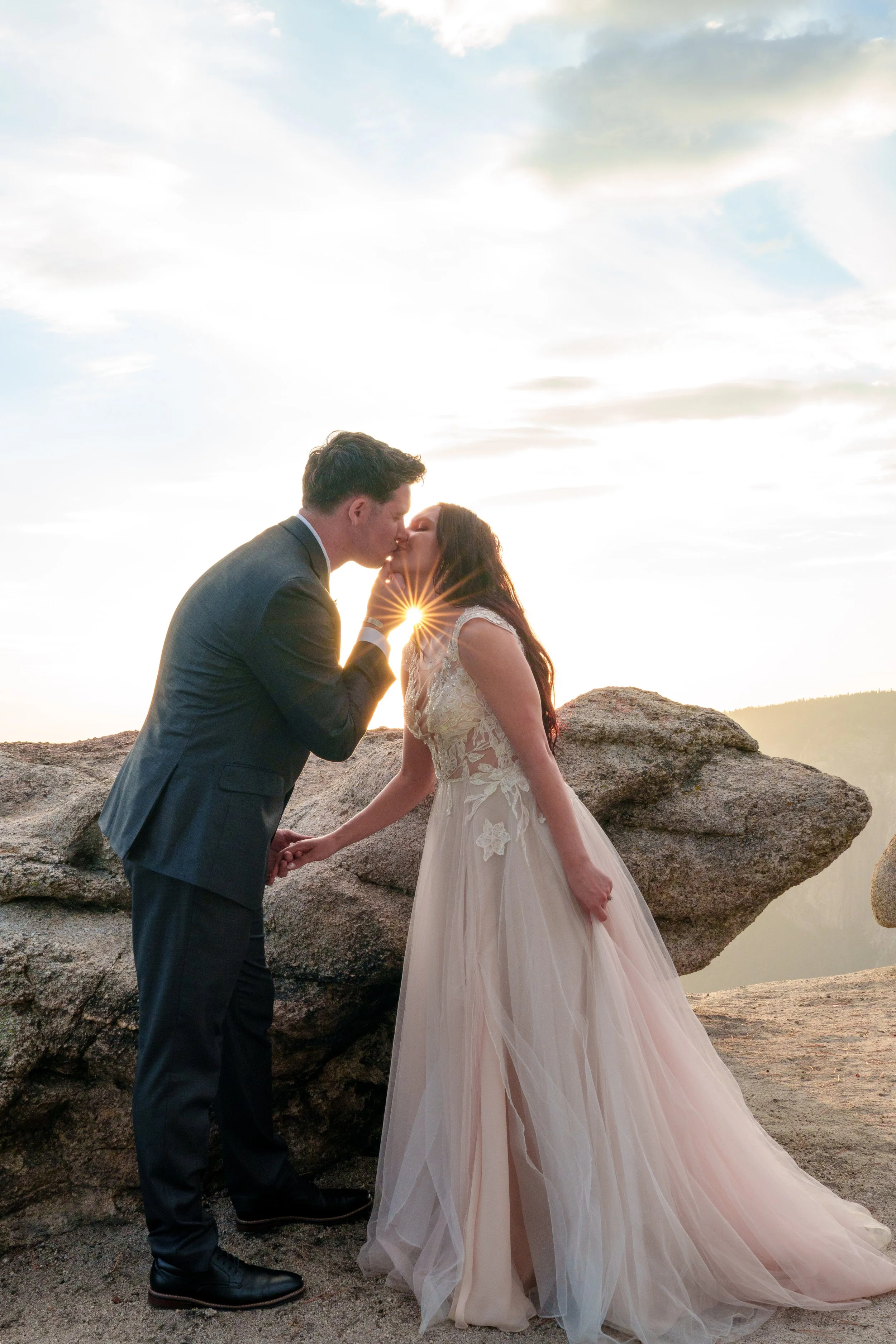 A couple in wedding attire sharing a kiss outdoors during sunset, with large rocks and a cloudy sky in the background. Yosemite Nation Park. Yosemite Elopement Photography. Yosemite Elopement Videography Taft Point