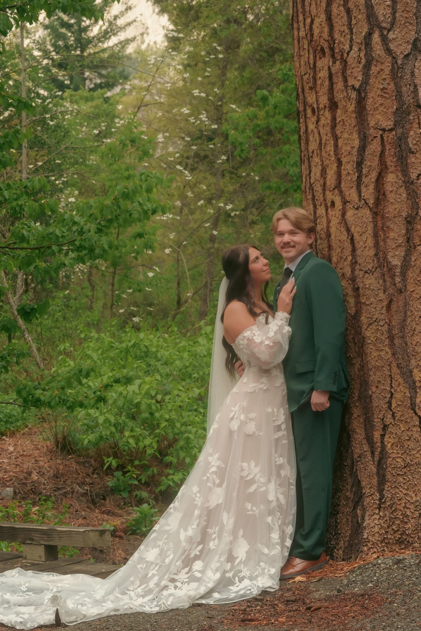 A bride and groom standing close together outside near a large tree, with greenery and foliage surrounding them. The bride is wearing a white floral lace wedding gown, and the groom is in a teal suit. They are looking at each other lovingly.