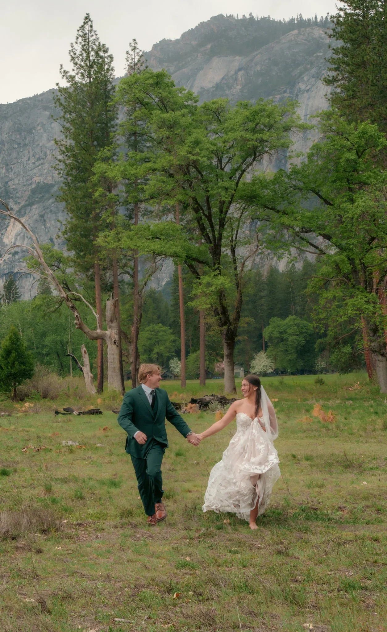 A bride and groom are holding hands and running through a grassy field, with mountains and trees in the background.