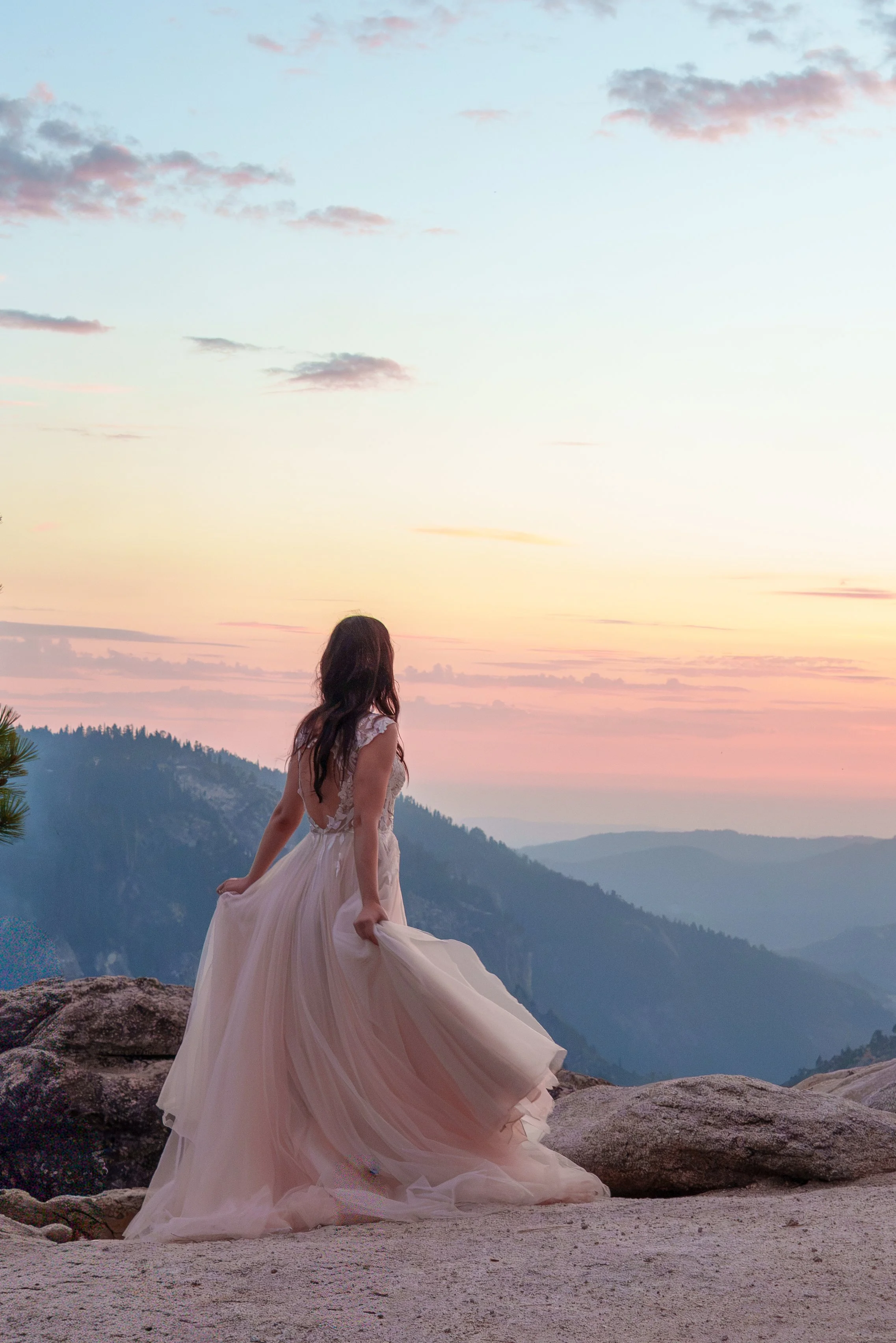 A woman in a flowing beige dress stands on rocks at sunset or sunrise, overlooking a mountainous landscape with a colorful sky and scattered clouds. Yosemite Nation Park. Yosemite Elopement Photography. Yosemite Elopement Videography Taft Point