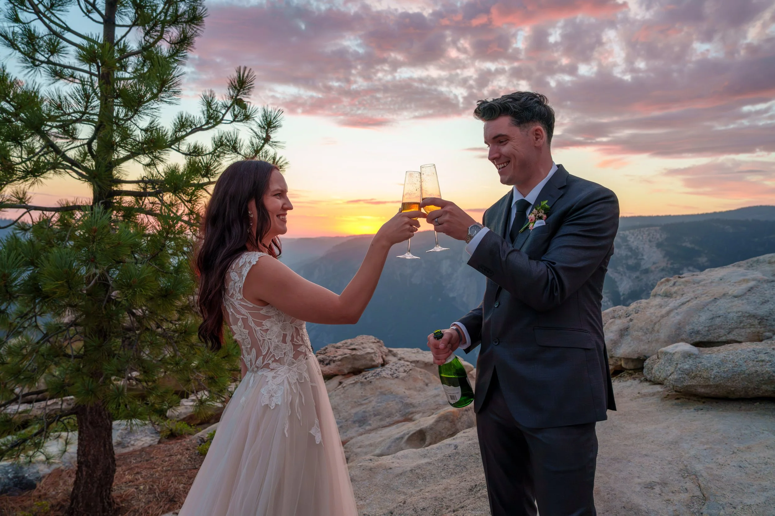 A couple dressed in wedding attire, smiling and toasting with champagne glasses at sunset on a mountain overlook, with a pine tree and rocky landscape in the background. Yosemite Nation Park. Yosemite Elopement Photography. Taft Point