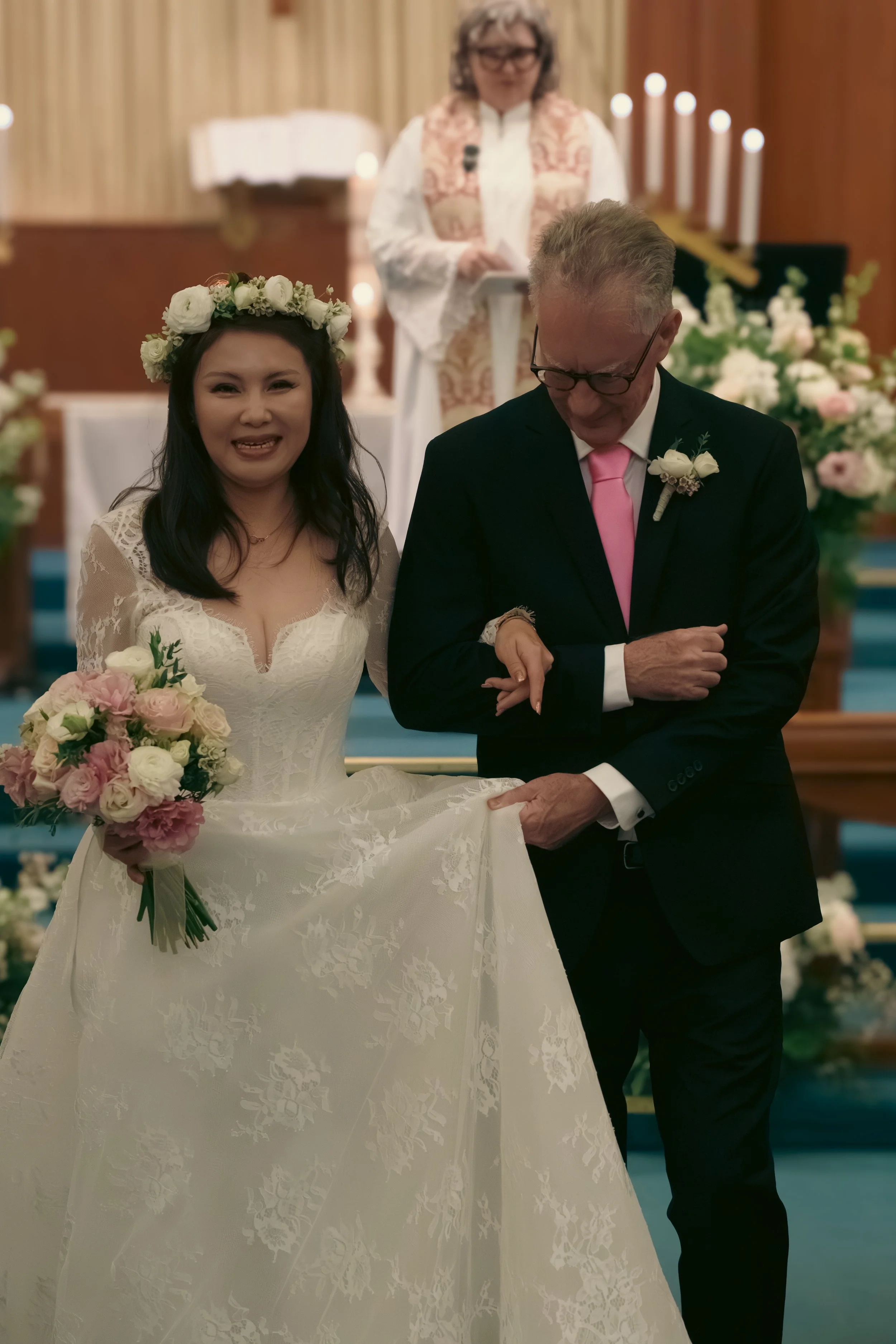 A bride in a white wedding dress holding a bouquet of pink and white flowers, smiling during her wedding ceremony with an older man in a dark suit, pink tie, and boutonniere, standing beside her. A woman officiant is in the background in front of an 
