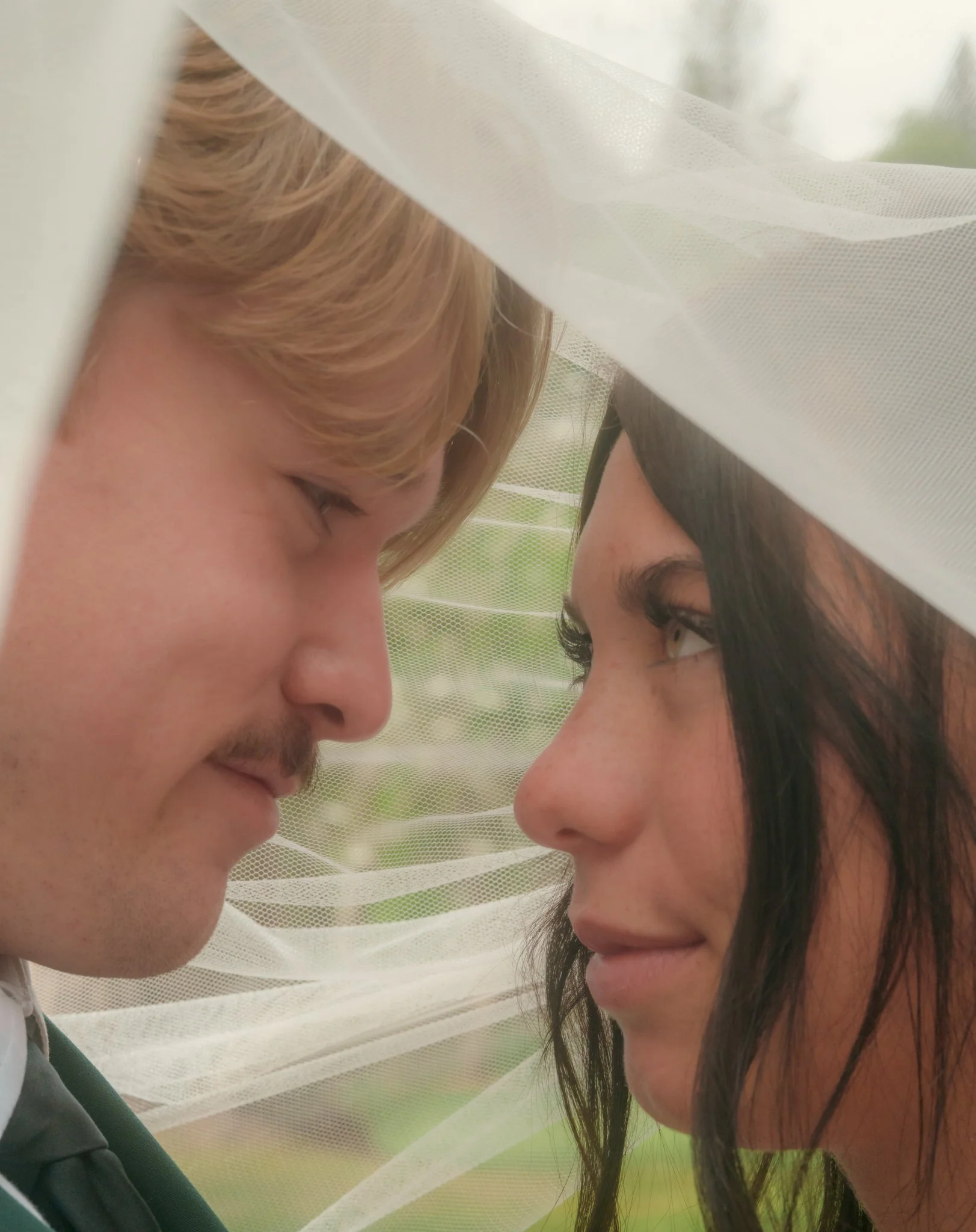 A close-up photo of a young man and woman with foreheads touching, eyes closed, under a sheer white canopy or veil outdoors.