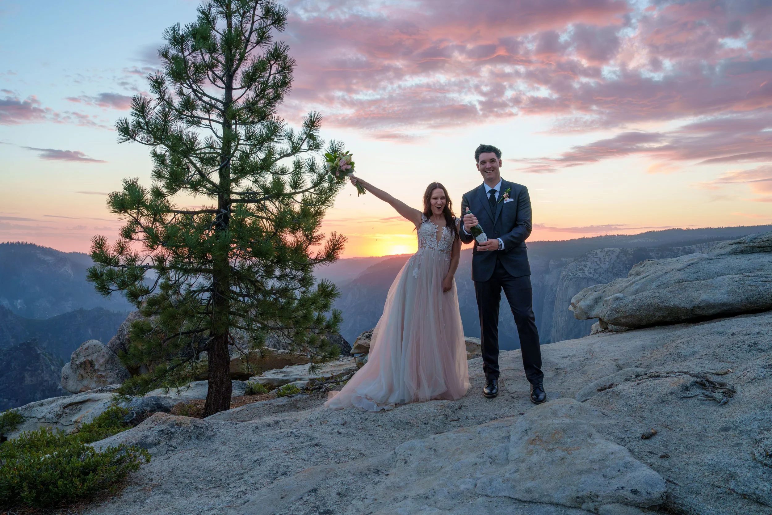 A happy couple dressed in formal attire, the woman in a pink dress and the man in a dark suit, celebrating on a rocky mountain Yosemite Nation Park. Yosemite Elopement Photography. Yosemite Elopement Videography Taft Point