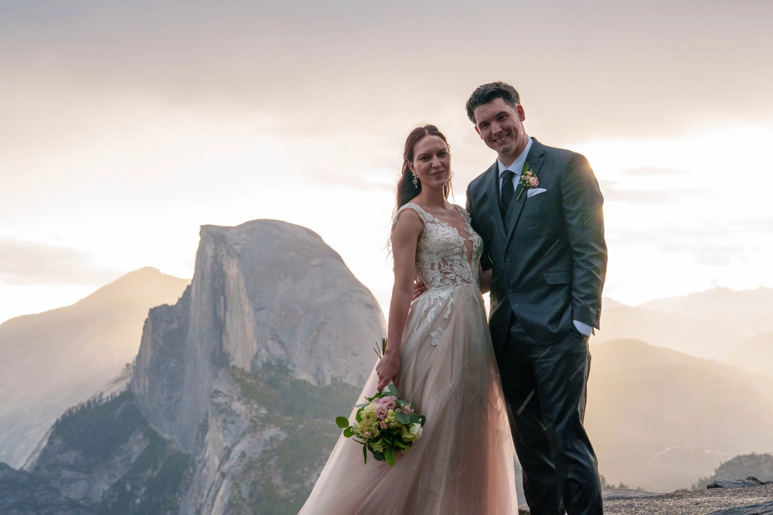 A newlywed couple stands on a mountain ridge at sunset, with a rocky peak and distant mountains in the background. The bride wears a lace wedding gown and holds a bouquet of flowers, and the groom wears a dark suit Yosemite Nation Park. 