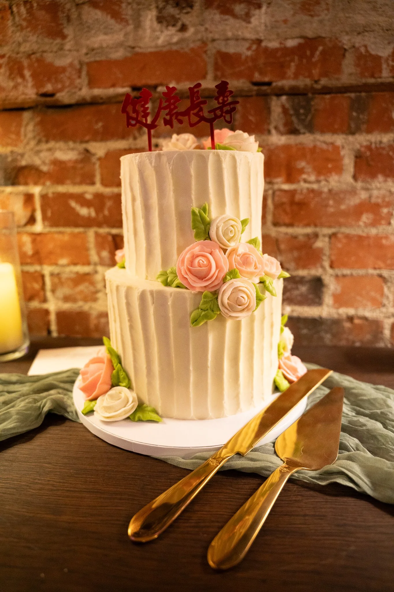 Two-tiered wedding cake decorated with buttercream flowers and green leaves, topped with a red Chinese character cake topper, placed on a wooden table with gold cake knife and server, against a brick wall background.