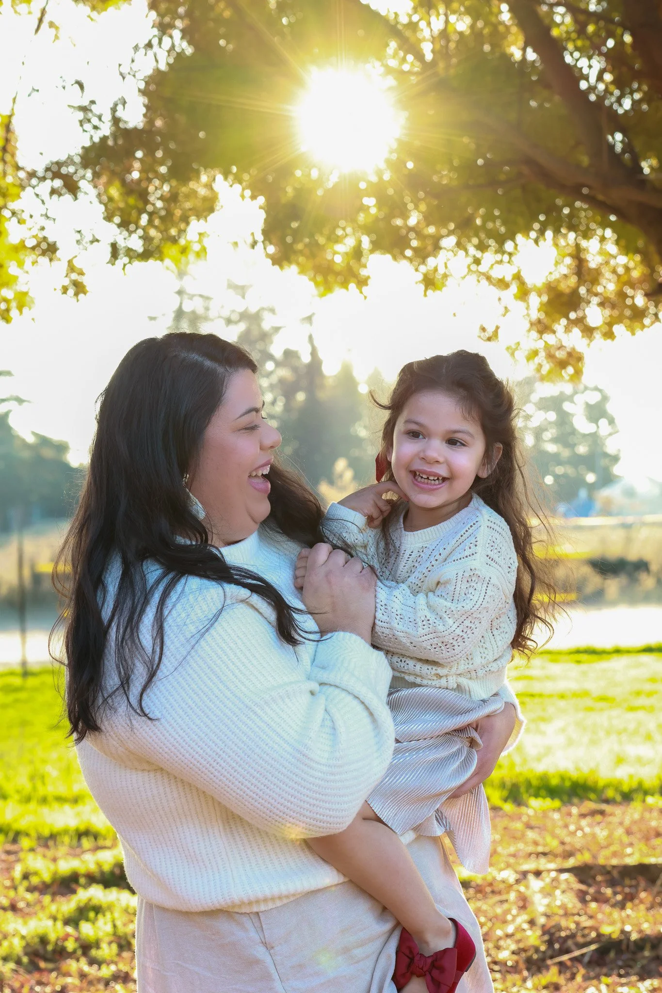 A woman holding a young girl outdoors during sunset, both smiling and enjoying each other's company.