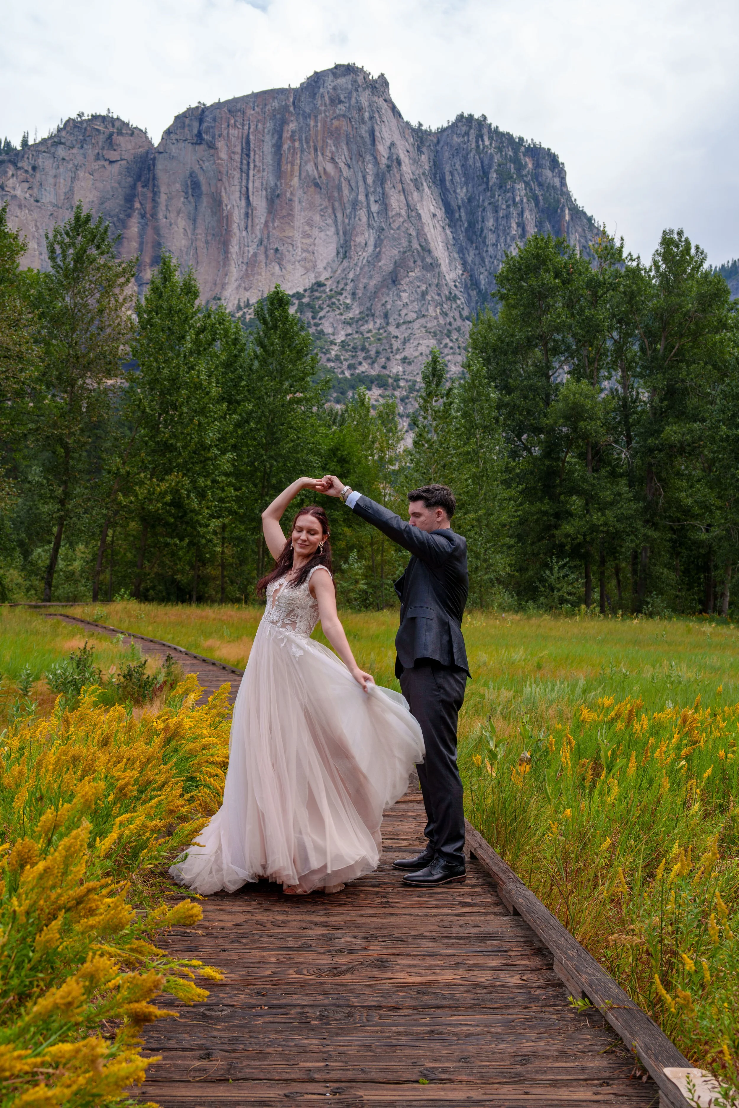 A bride and groom dancing on a wooden path surrounded by yellow flowers and green trees in front of a mountain landscape. Yosemite Nation Park. Yosemite Elopement Photography. Yosemite Elopement Videography Cathedral Beach