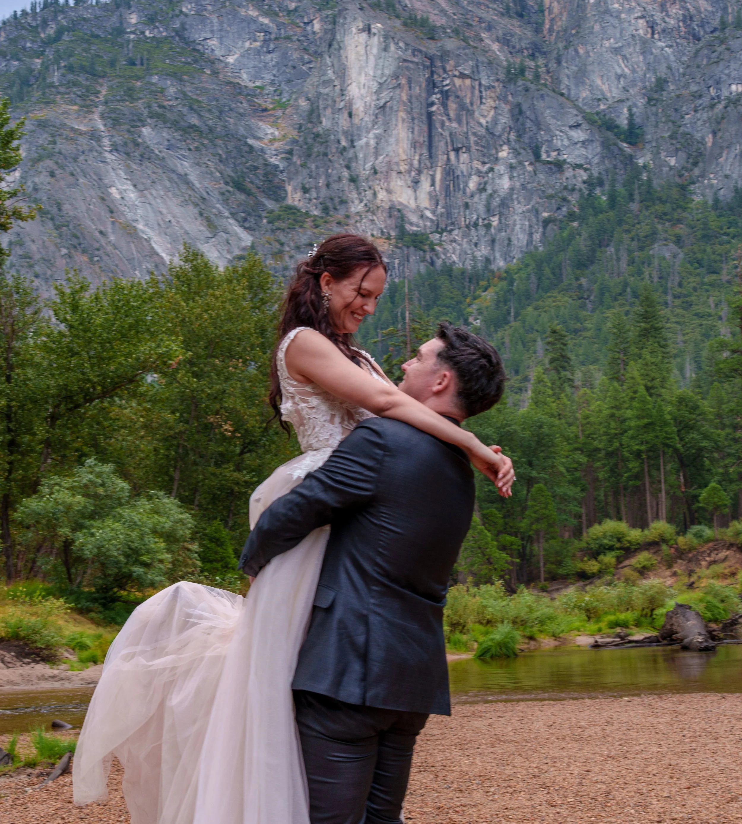 A man in a suit lifting a woman in a wedding dress outdoors near a river, with tall rocky mountains and green trees in the background. Yosemite Nation Park. Yosemite Elopement Photography. Yosemite Elopement Videography Cathedral Beach
