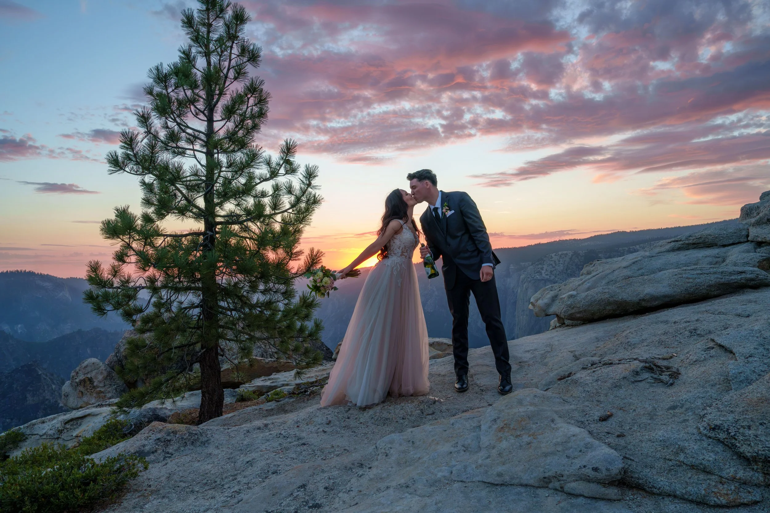 A couple dressed in wedding attire sharing a kiss on a rocky mountain overlook at sunset, with a pine tree in the foreground and colorful clouds in the sky. Yosemite Nation Park. Yosemite Elopement Photography. Yosemite Elopement Videography
