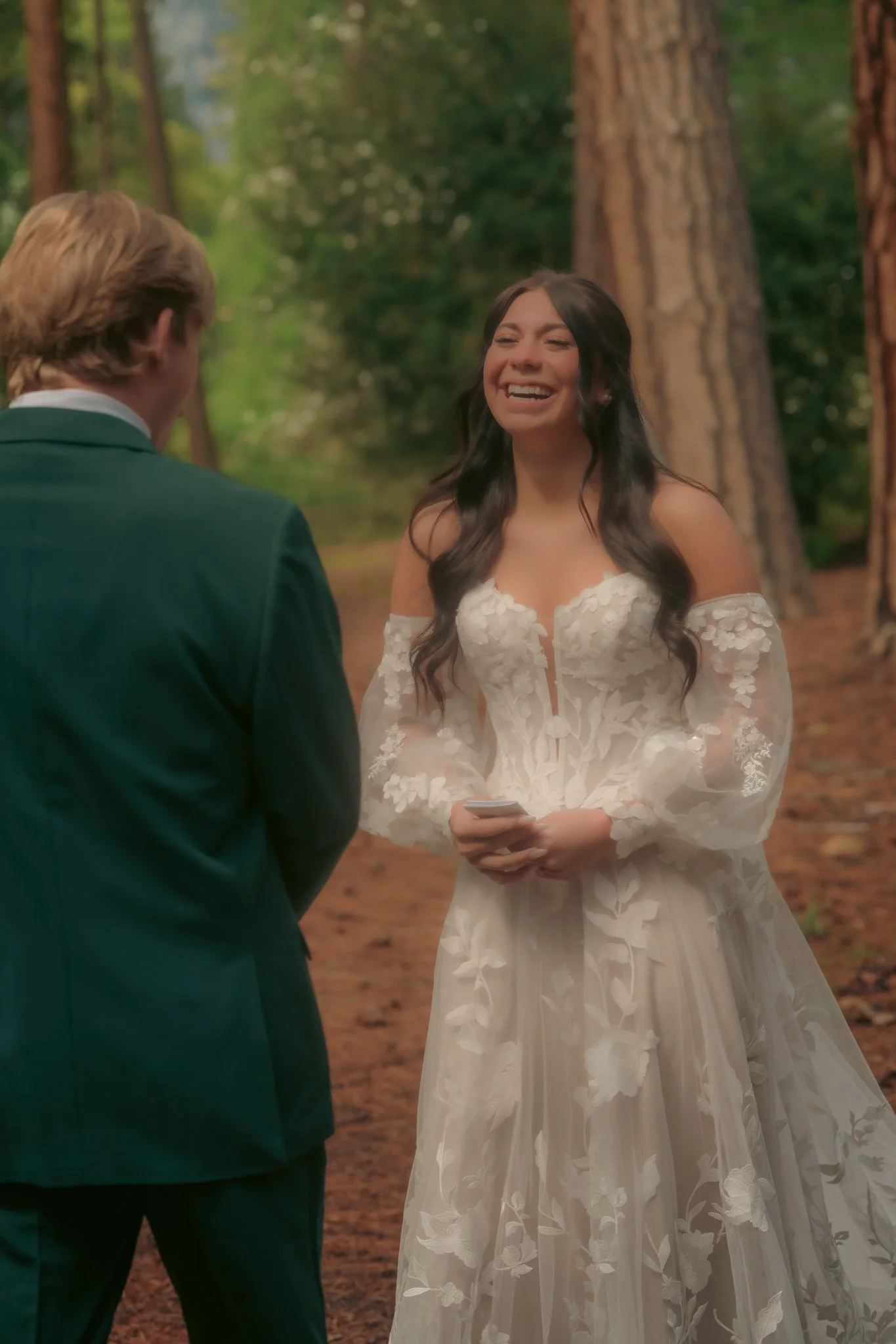 A woman in a white floral wedding dress smiling and holding a small card during an outdoor wedding ceremony in a forested area.