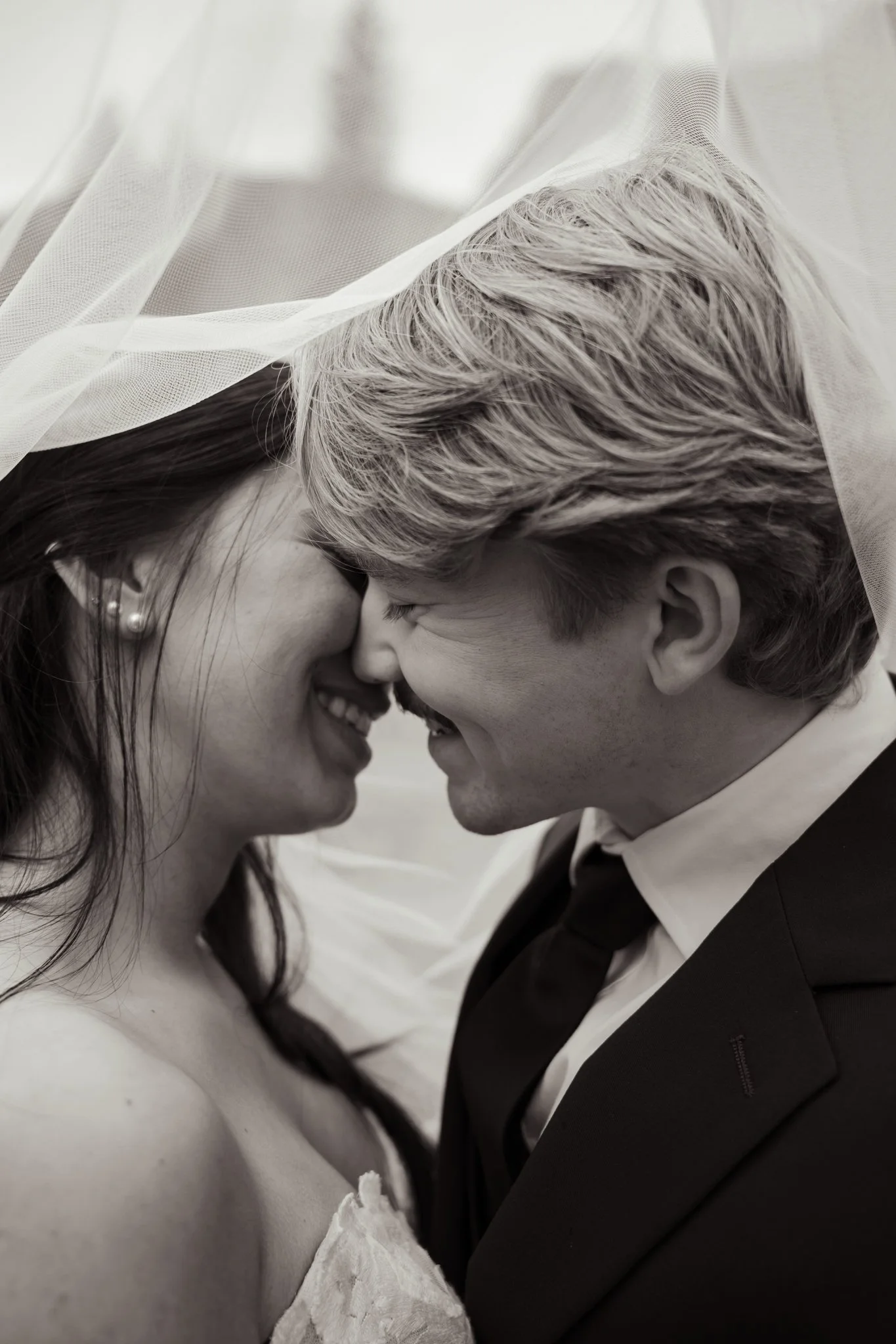 A black-and-white close-up photo of a smiling couple with their foreheads touching, partially covered by a translucent veil, dressed in wedding attire.