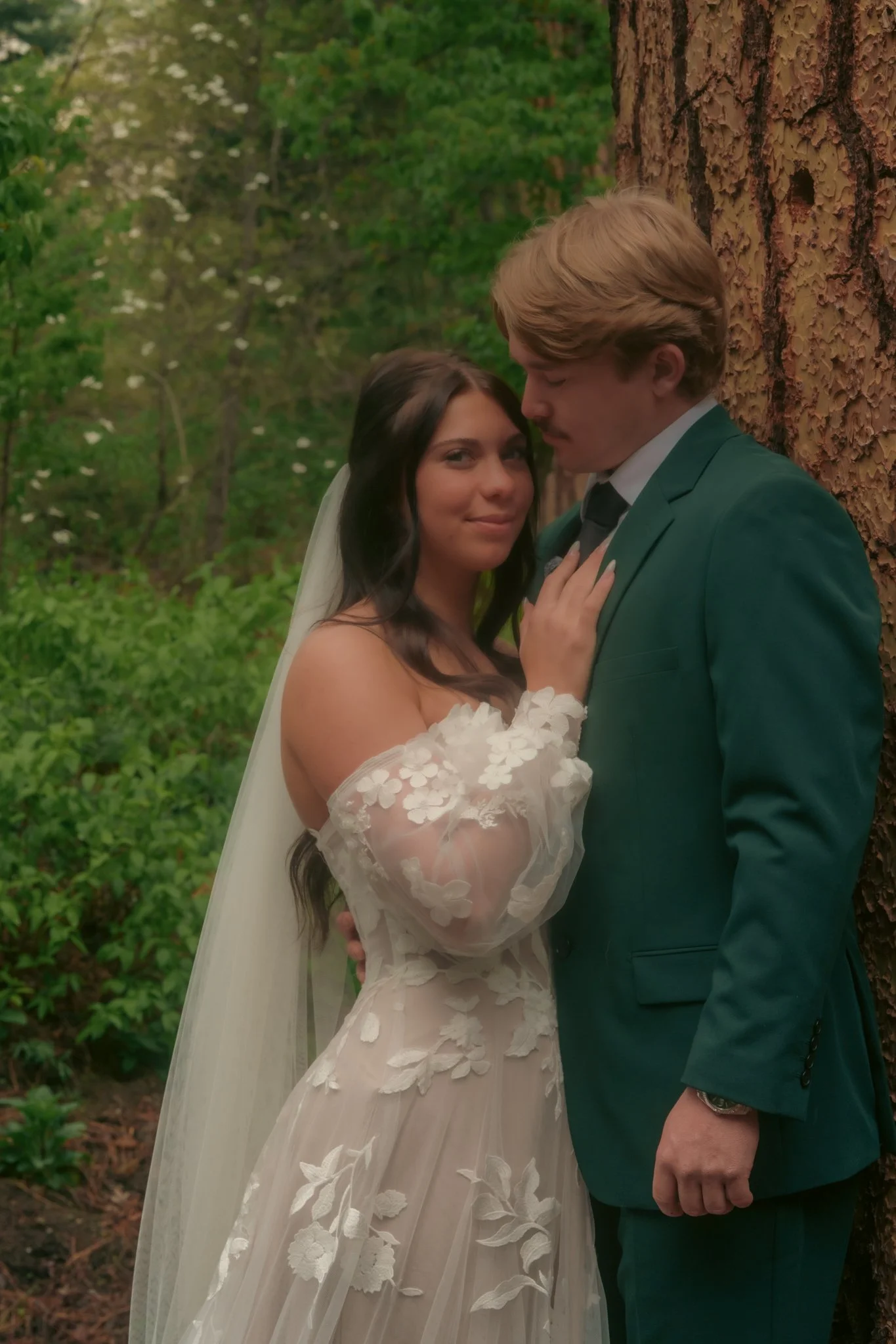 A bride and groom are standing close together outdoors, with the bride leaning against a tree and the groom gently pressing his forehead to her head. The bride is wearing a white floral lace wedding dress and a veil, while the groom is in a green sui