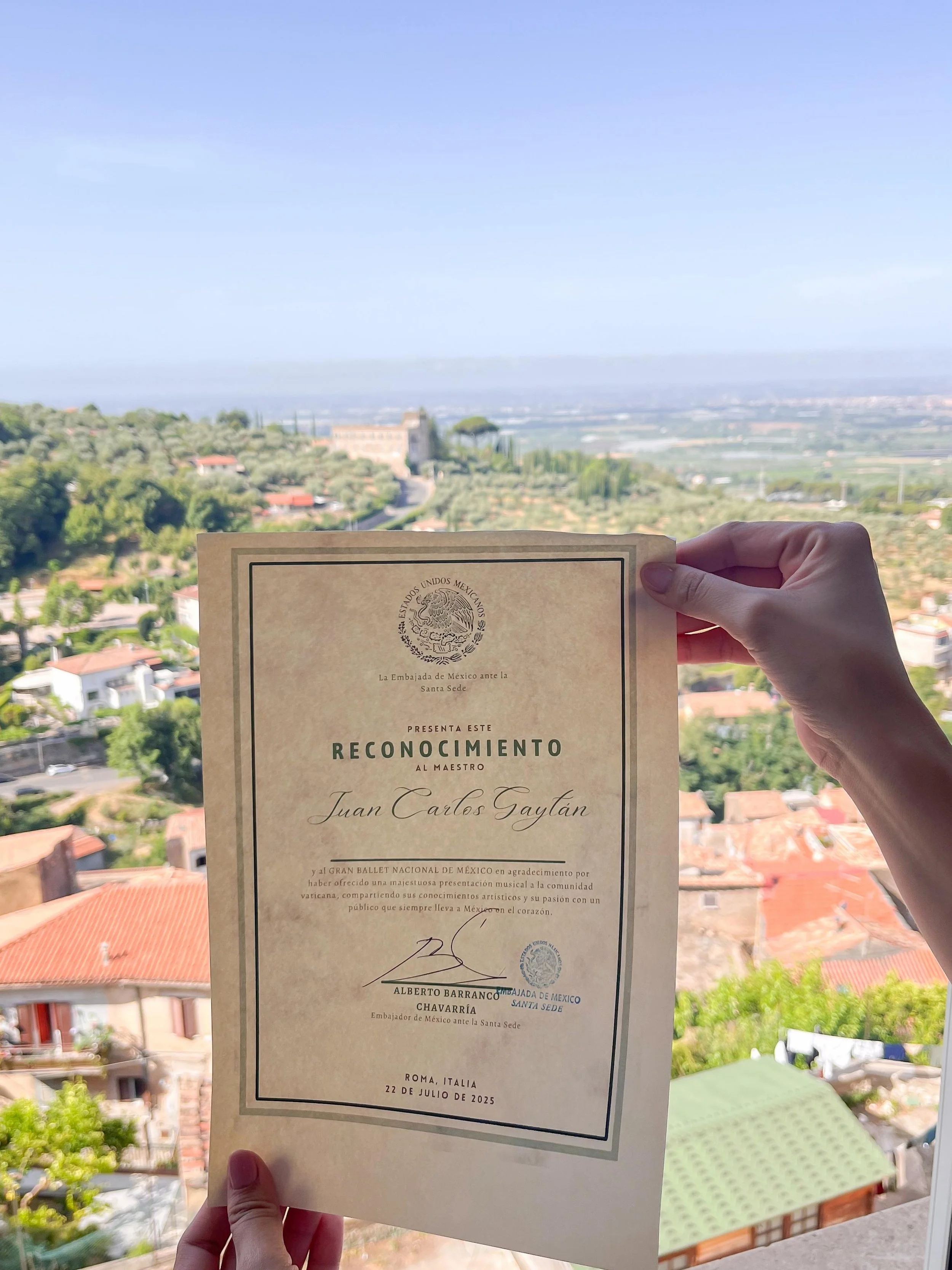 Person holding a certificate from the Embassy of Mexico in front of a scenic view of a hilly landscape with houses, trees, and fields.