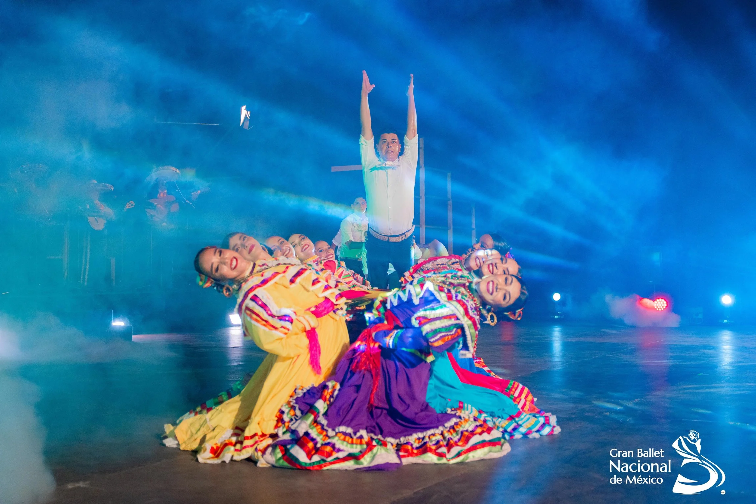 Performance of dancers in colorful traditional dresses on stage with blue lighting and fog, with a male performer standing with arms raised, at Gran Ballet Nacional de México.