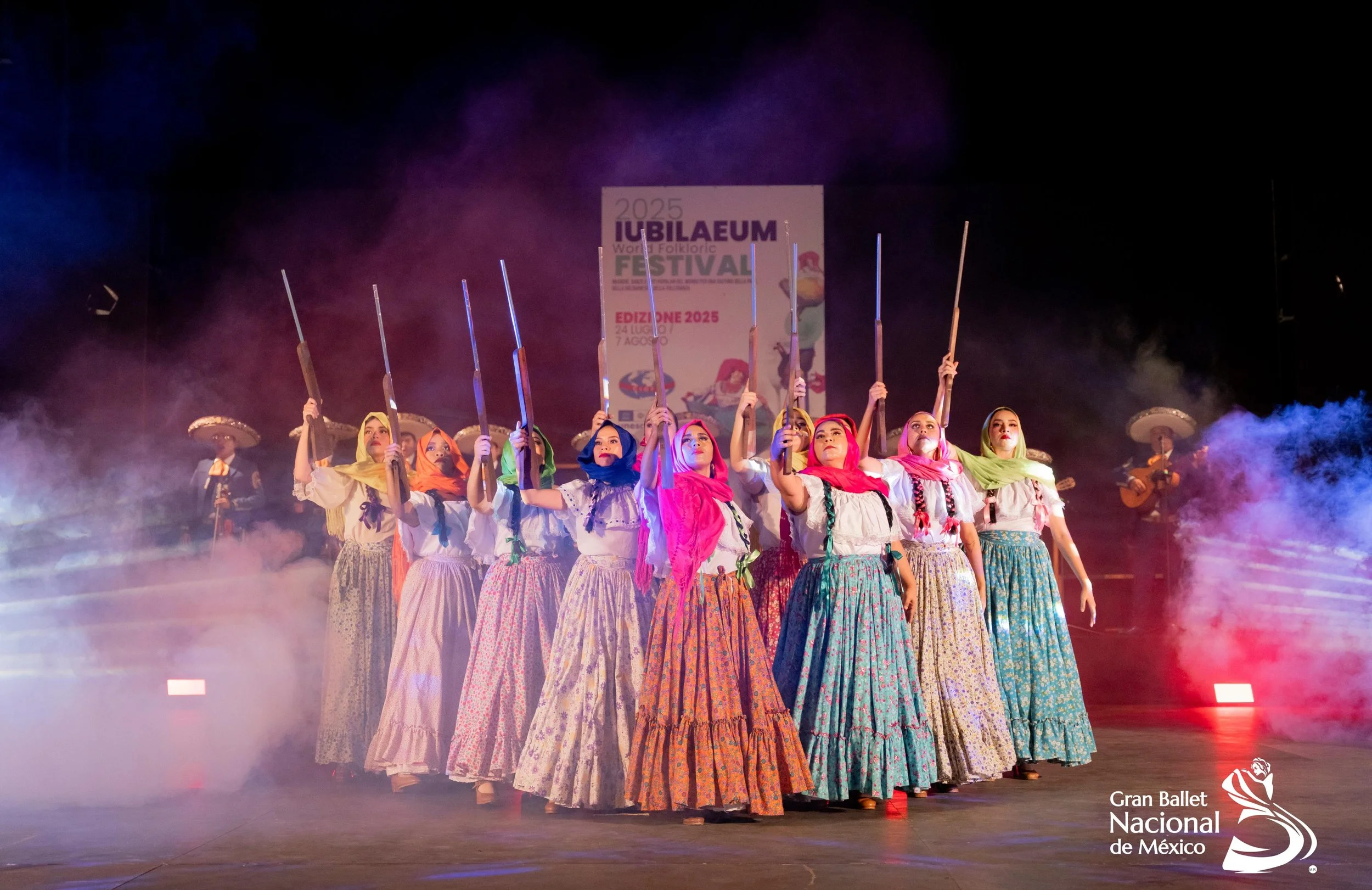 Group of female dancers in colorful traditional Mexican costumes with headscarves, holding rifles in a choreographed performance on stage during a cultural event, with musicians in the background and a foggy atmosphere.
