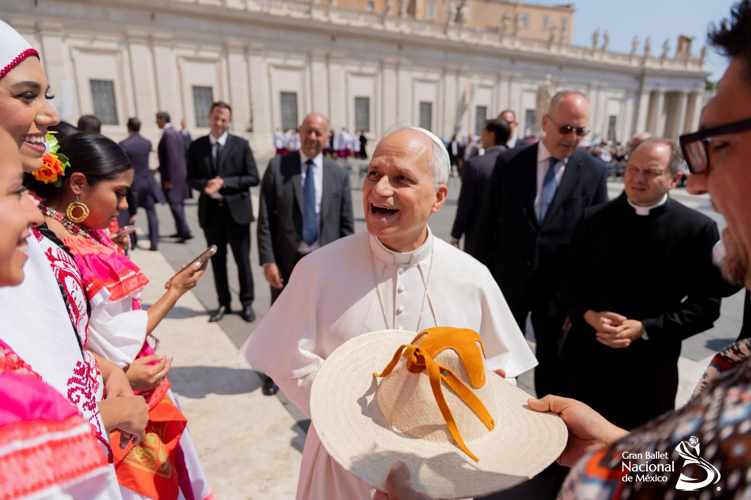 Pope Francis smiling and holding a sombrero with a ribbon, surrounded by people, some in colorful traditional Mexican clothing, in front of a historical building, during an outdoor event.
