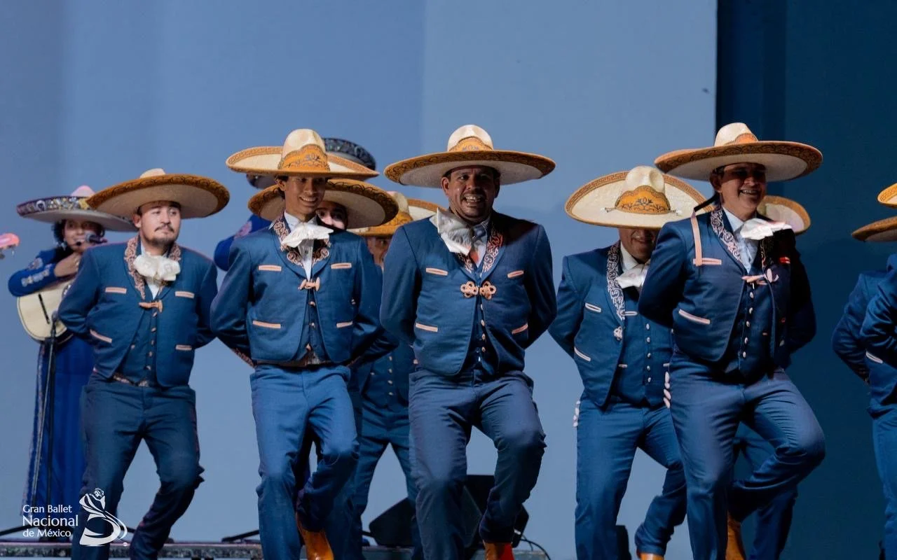 A group of dancers in traditional Mexican attire wearing blue suits with pink accents and large straw sombreros, performing on stage with a blue backdrop. The dancers are part of the Gran Ballet Nacional de México.