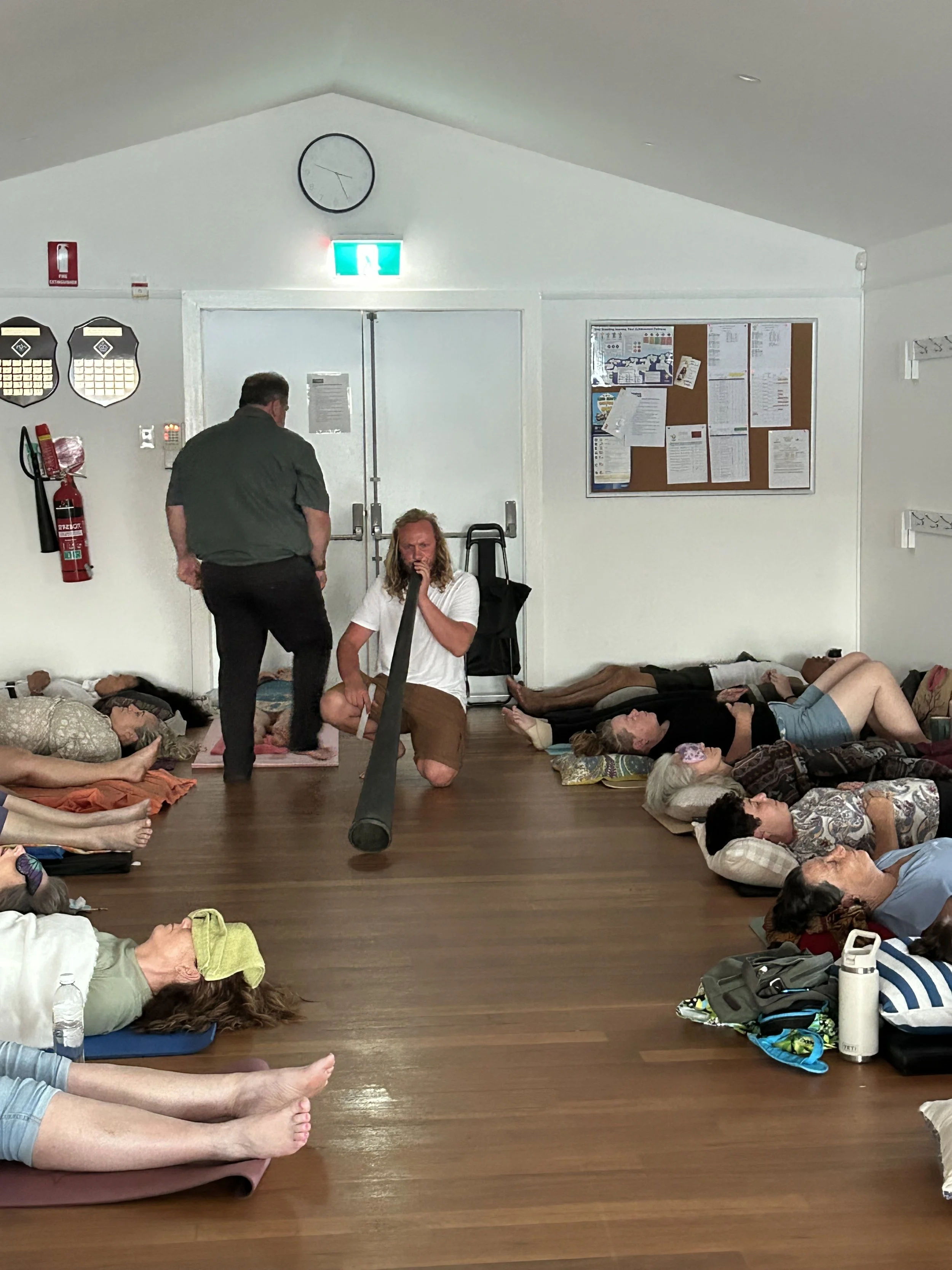 People lying down on the floor in a room, some resting and some in a meditative or relaxation pose, with bags and personal items around them, and two men sitting and talking near the door.