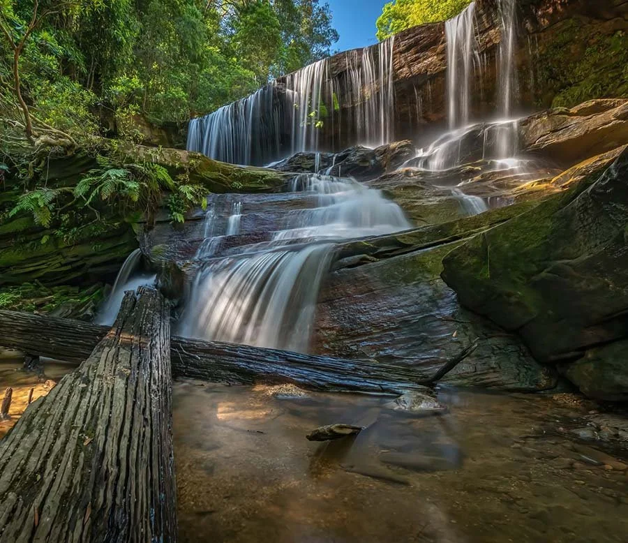 A multi-tiered waterfall flowing over rocks in a lush green forest, with a fallen log in the foreground and sunlight filtering through the trees.