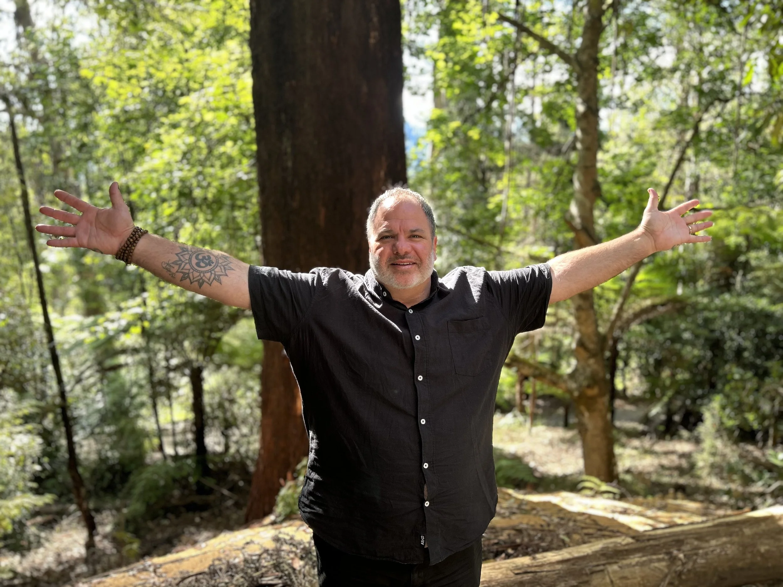 A man stands outdoors in a forest with green trees in the background, arms outstretched, smiling, wearing a black shirt.