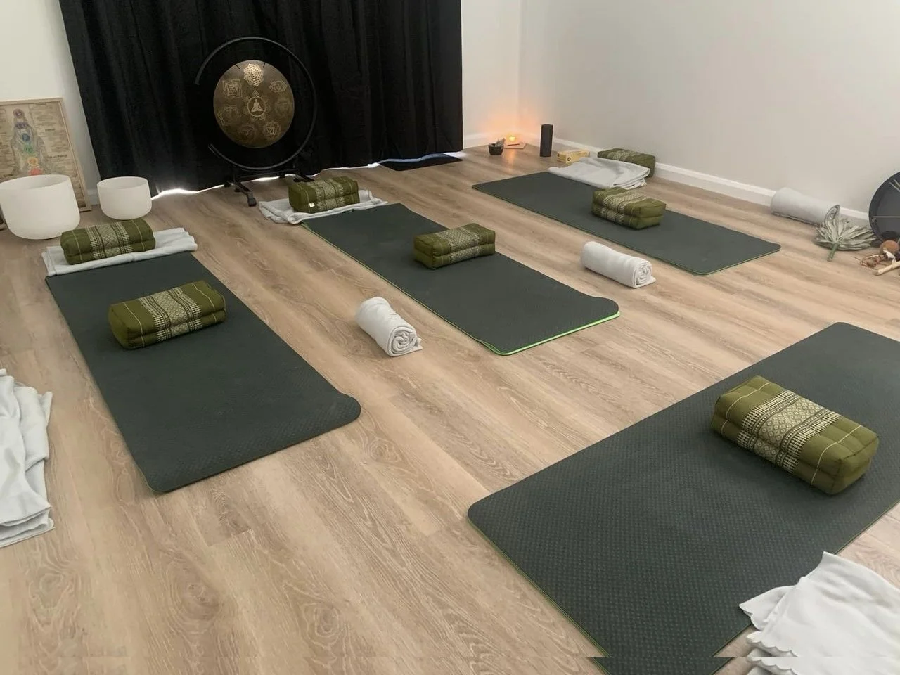 Yoga mats with rolled towels and blankets arranged in a meditation or yoga class setup on a wooden floor, with some objects and decor in the background.