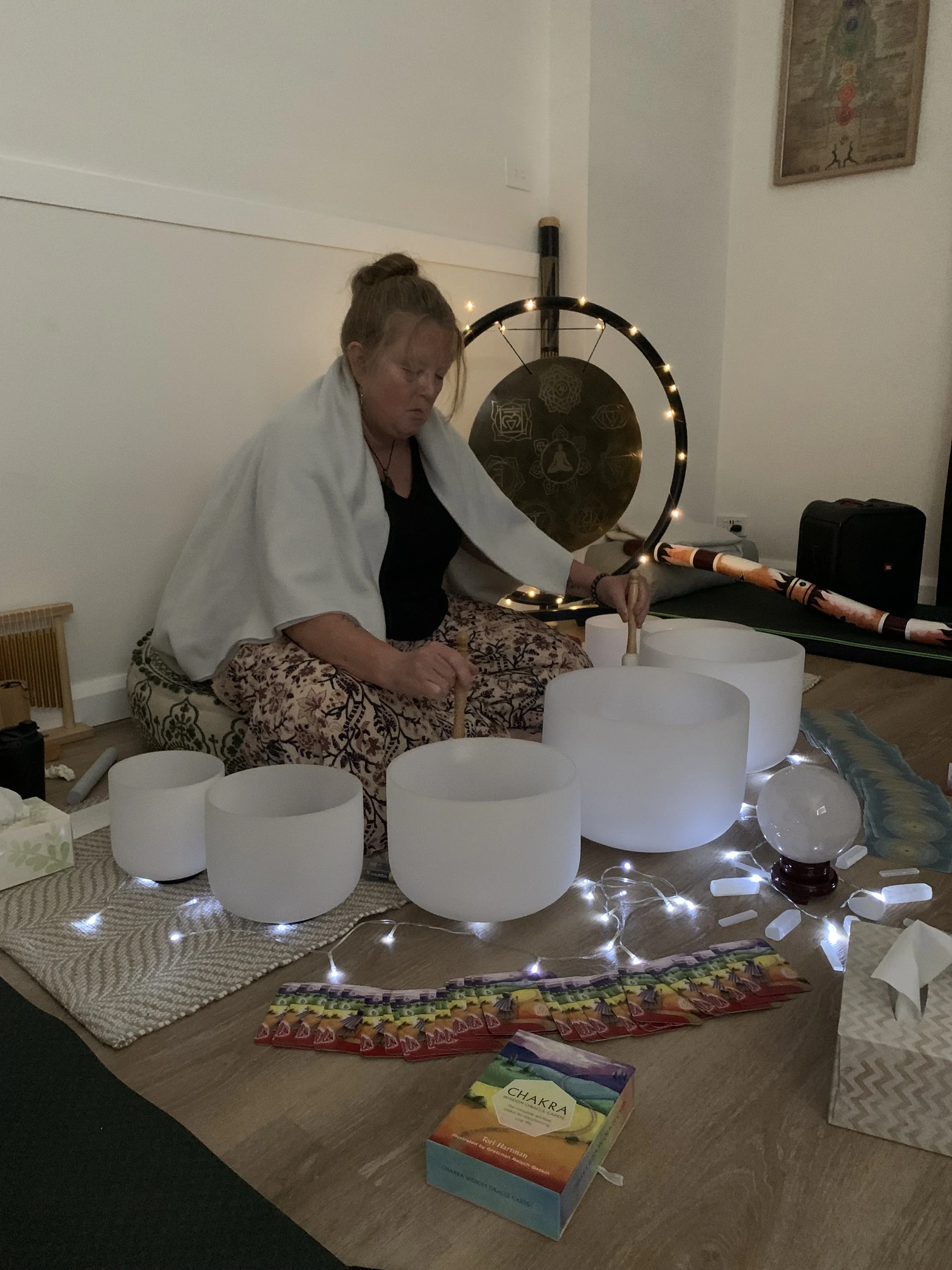 A woman with braided hair, sitting cross-legged on a patterned cushion, playing crystal singing bowls surrounded by fairy lights, oracle cards, and a healing crystal.