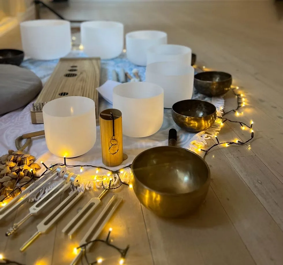 Set of singing bowls and mallets arranged on a wooden floor with decorative lights and a cloth backdrop, used for sound therapy or meditation.