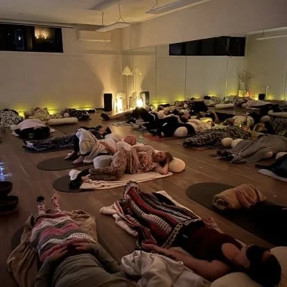 People participating in a group meditation or relaxation session in a dimly lit studio, lying on mats with blankets and pillows.