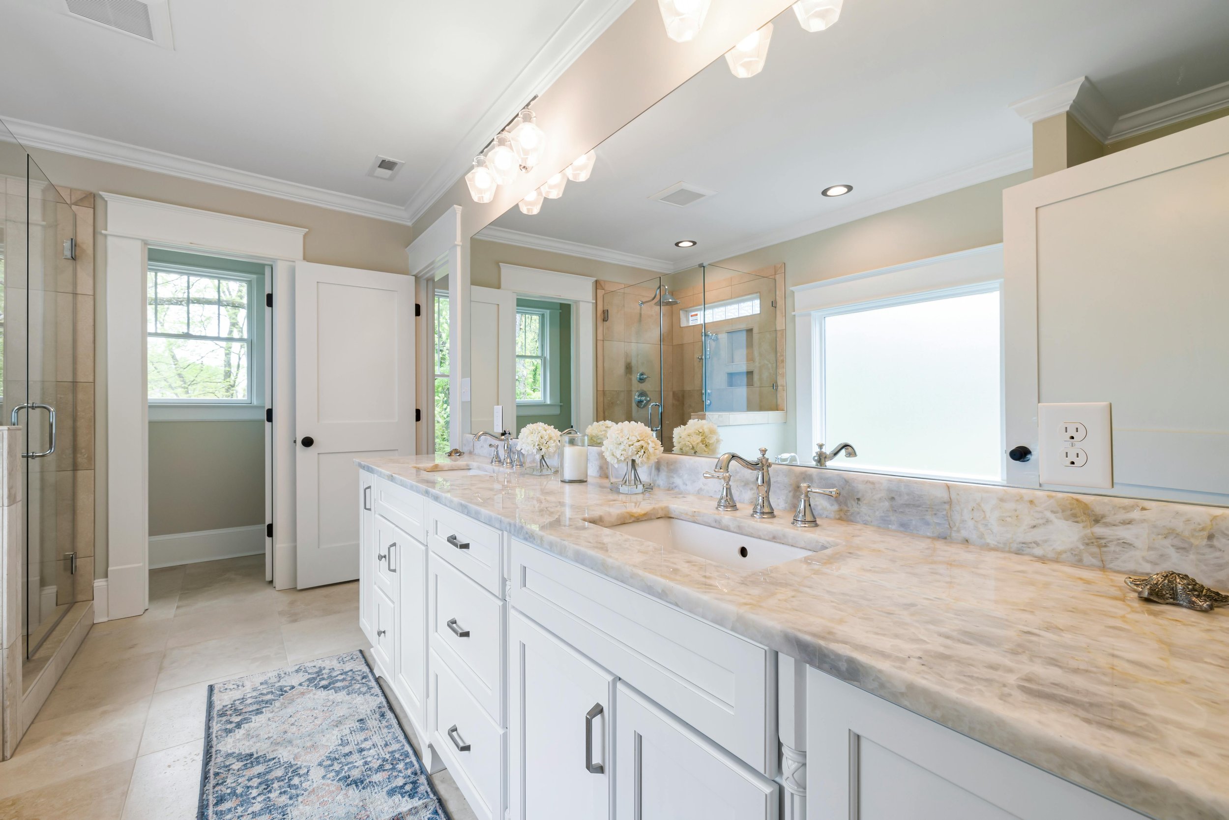 A bright, modern bathroom featuring a double sink vanity with a marble countertop, floral arrangements, a glass-enclosed shower, and large windows providing natural light.