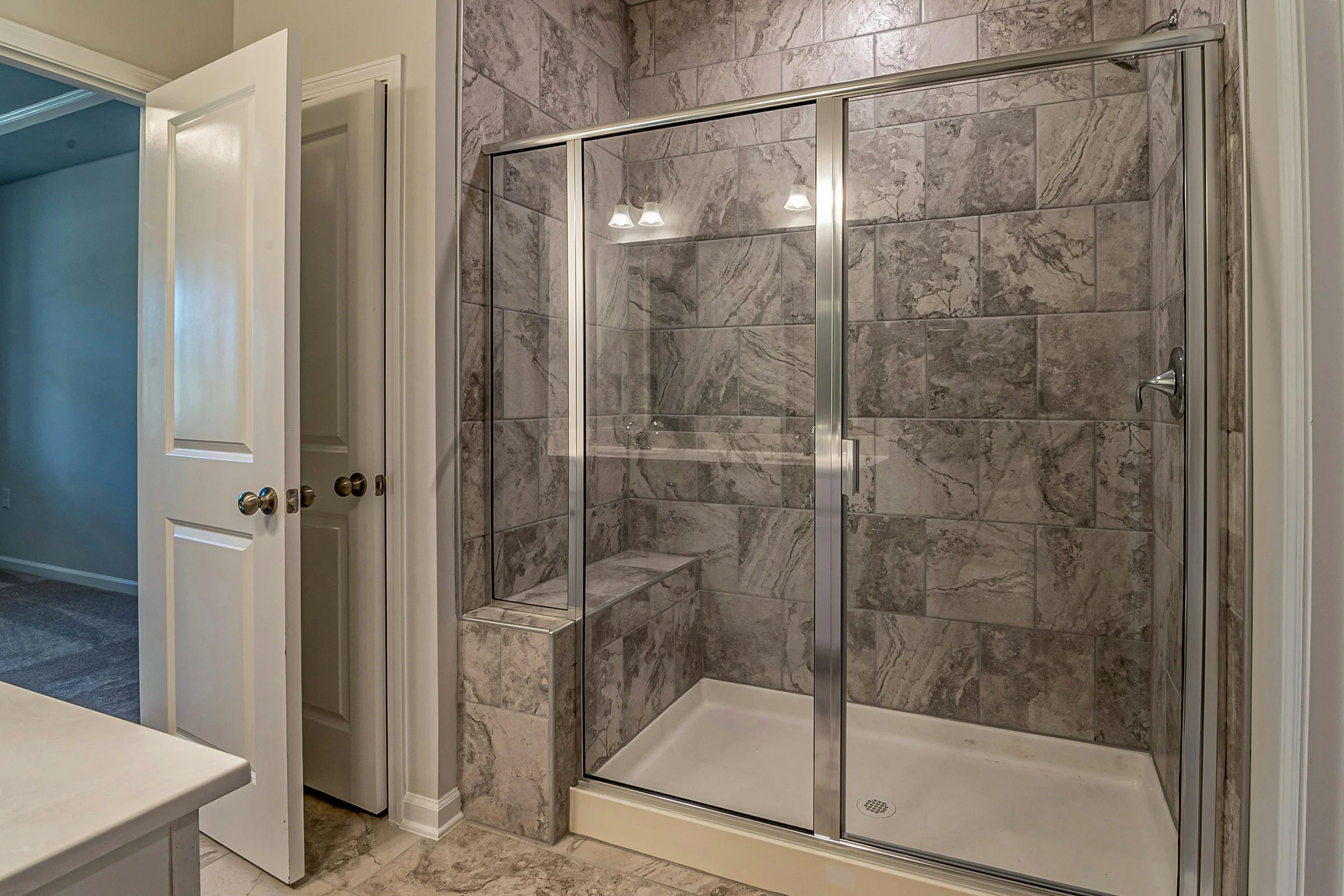 A tiled bathroom shower with sliding glass doors, built-in bench, and overhead lighting, with an open door leading to a carpeted room.