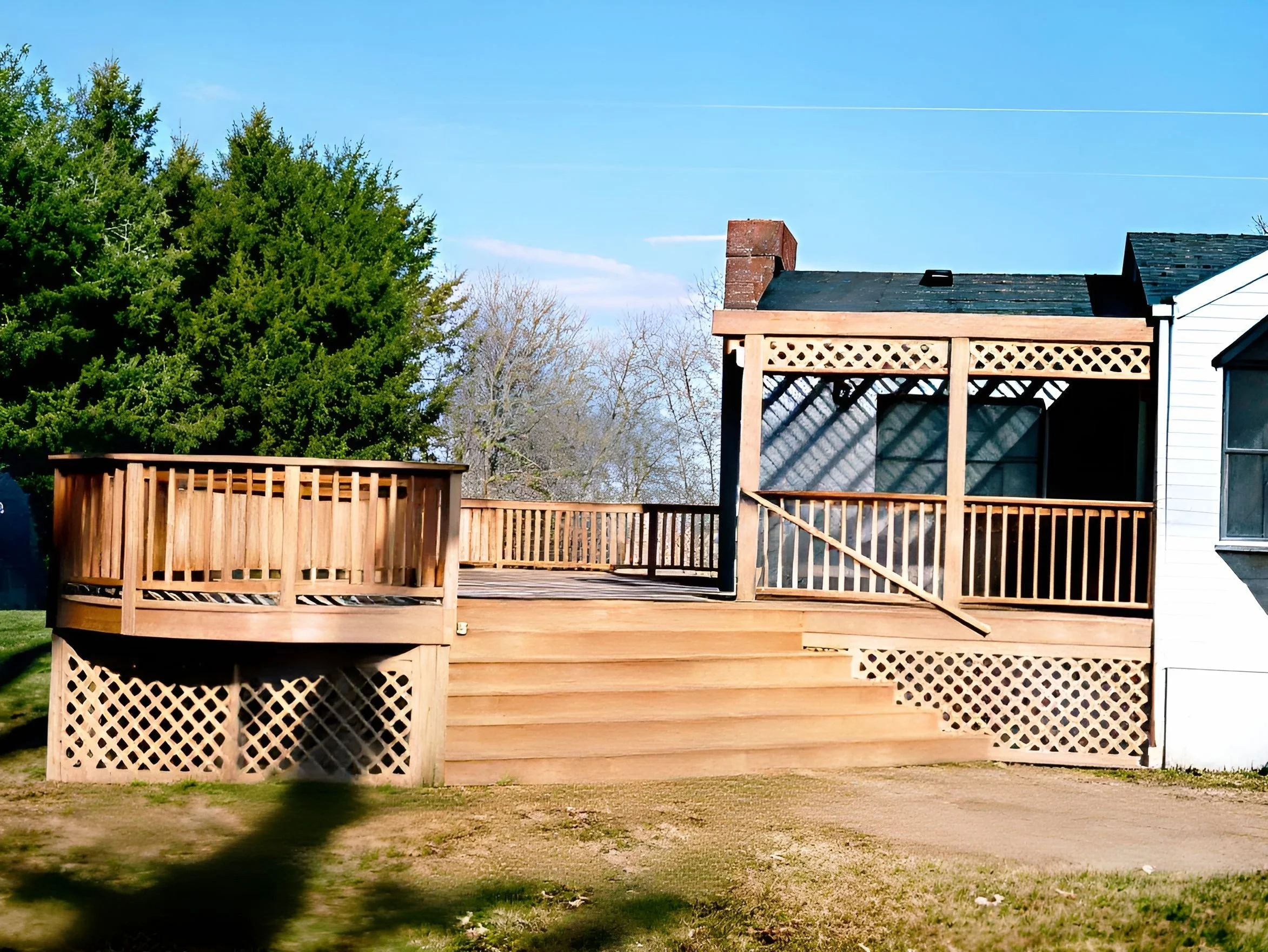 Newly constructed wooden deck with railings and lattice skirting, stairs leading up to it, attached to a white house on the right, with green trees and clear blue sky in the background.
